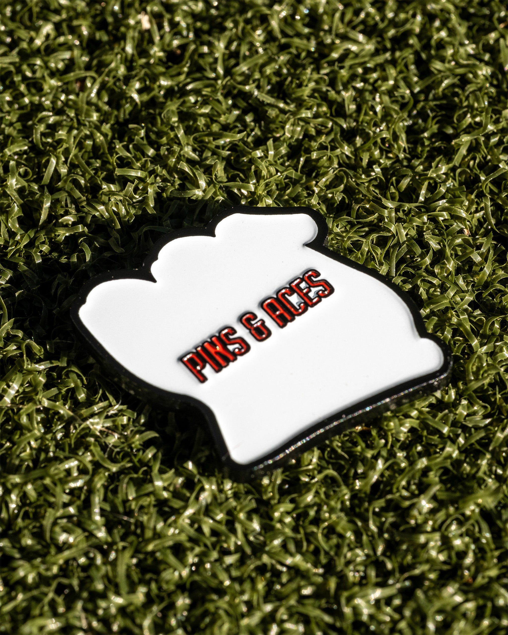 White ball marker with "Pins & Aces" text on a green turf background. Ideal for golf enthusiasts.