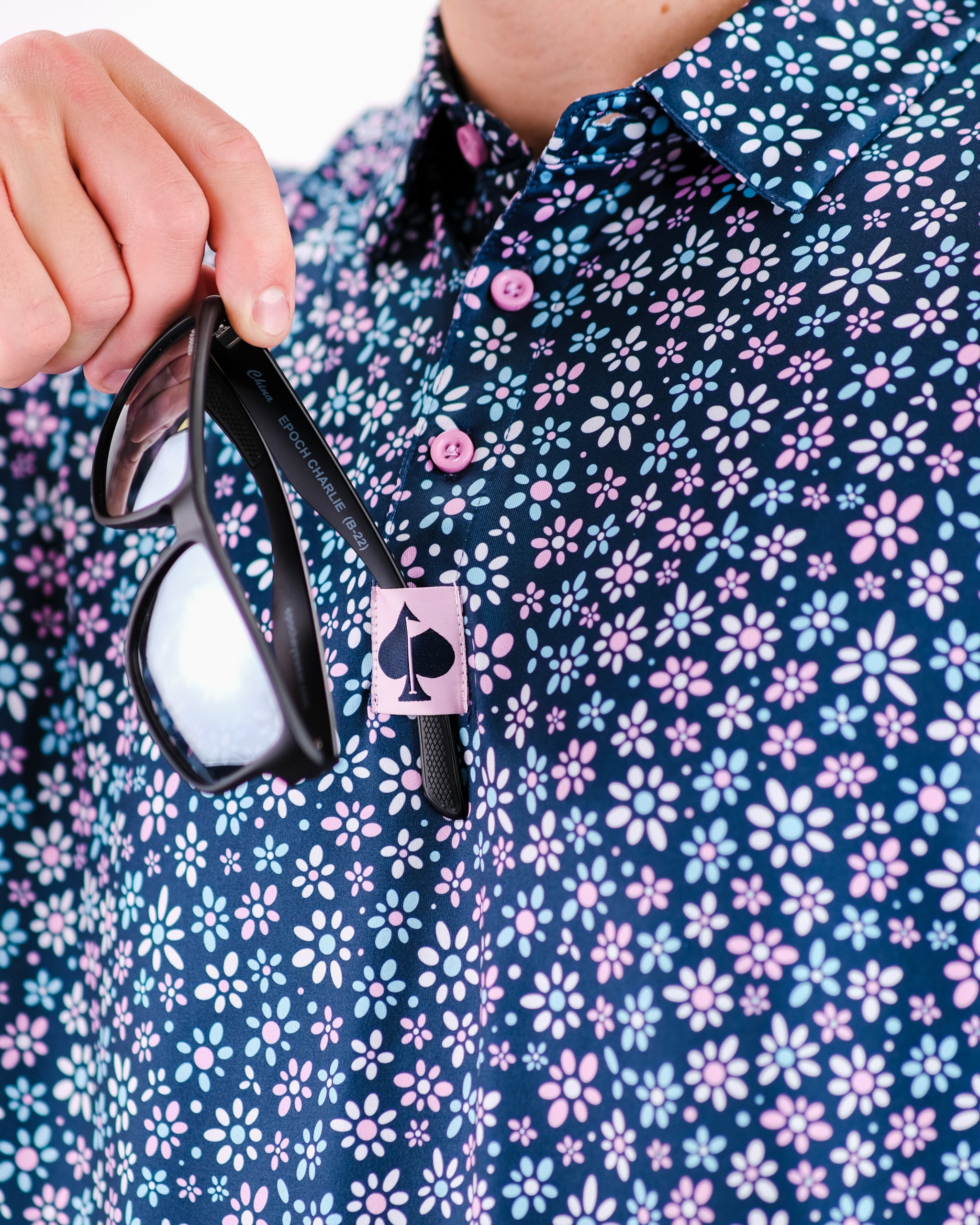 Men's floral golf shirt in navy with pink and blue daisies, featuring a stylish collar and sunglasses hanging from the button.