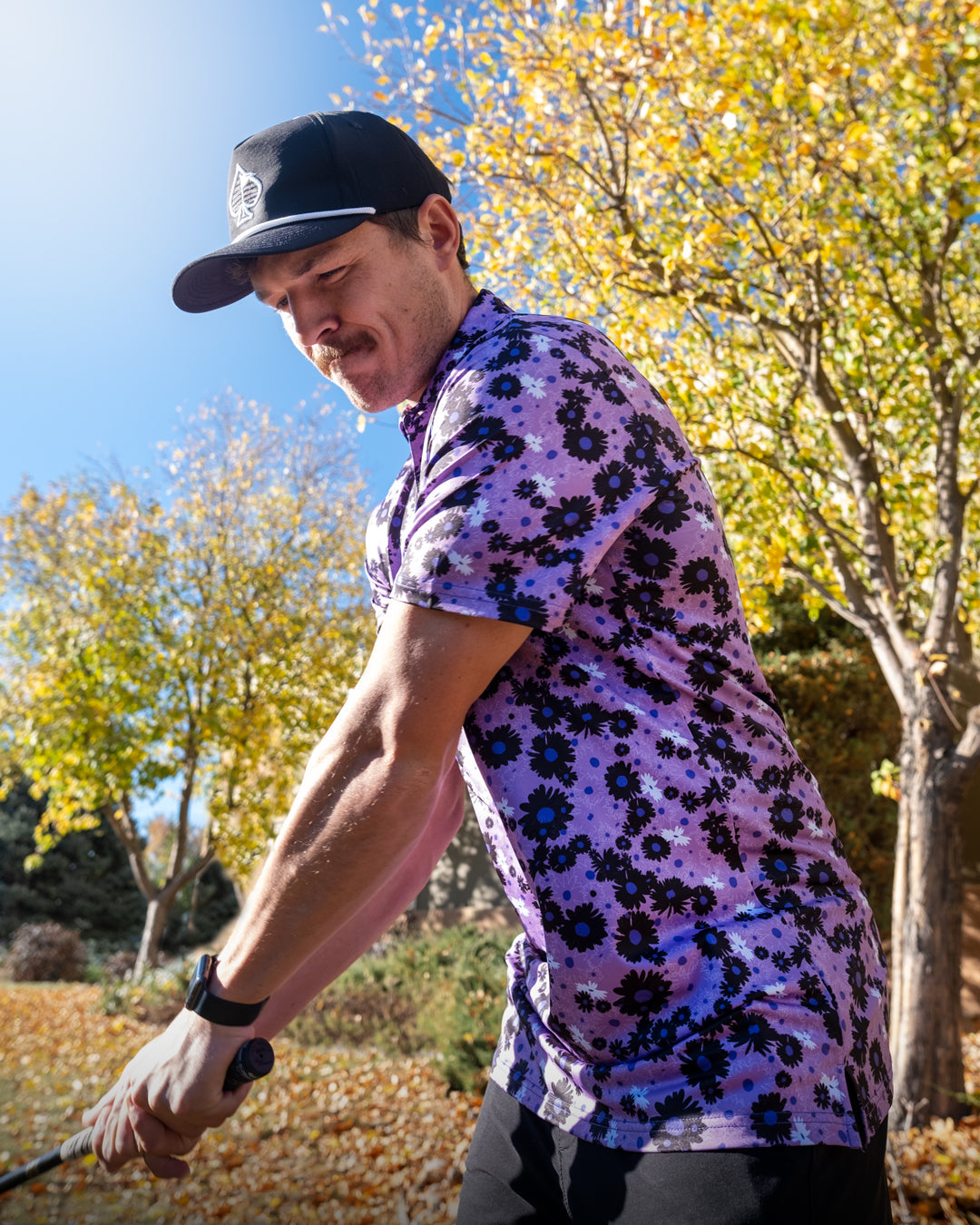 A golfer swings a club while wearing a vibrant purple floral shirt against a backdrop of autumn trees.