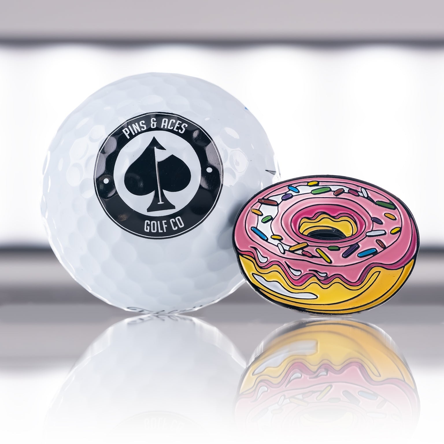 Colorful donut-shaped ball marker next to a golf ball featuring the Pins and Aces logo, perfect for stylish golfers.