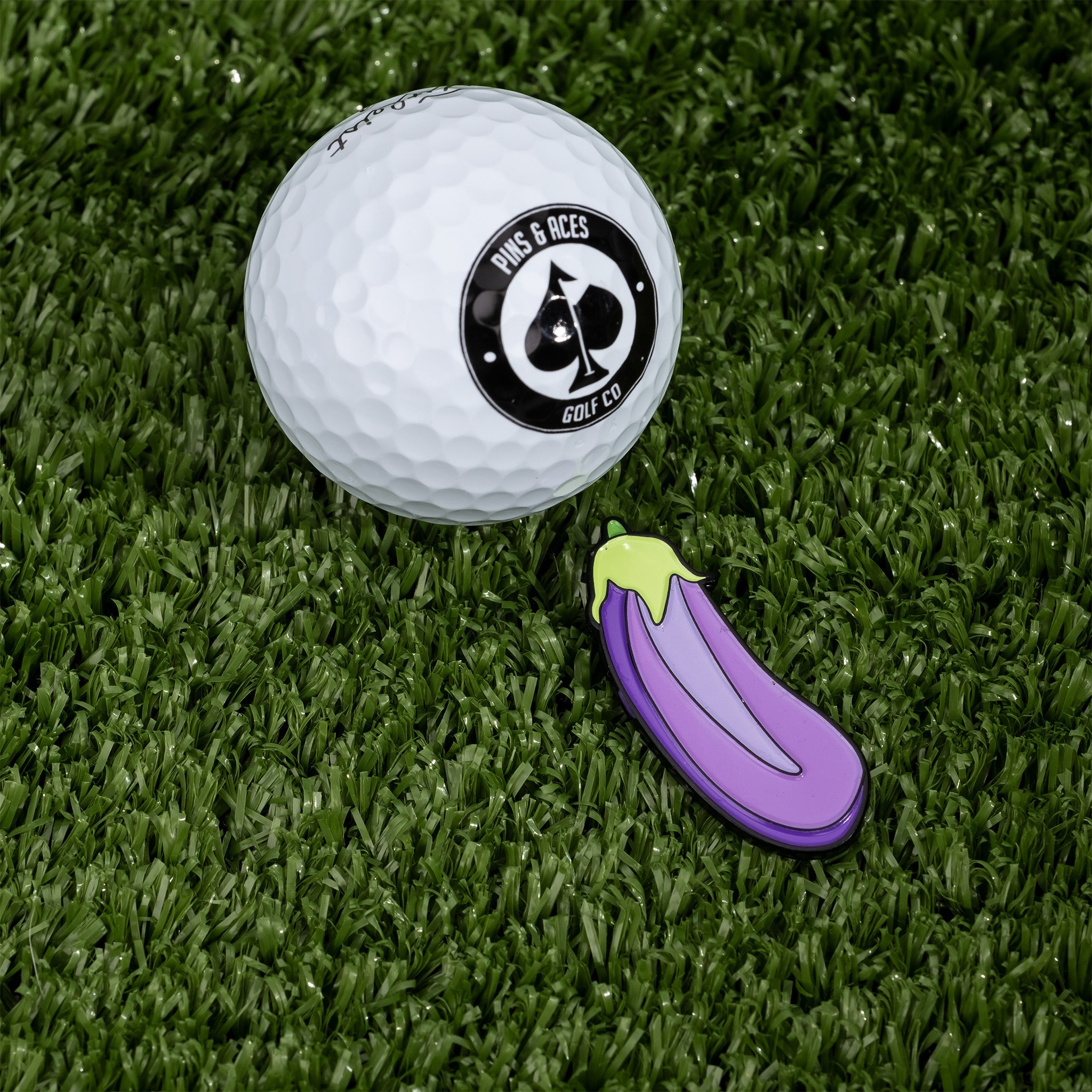 Eggplant-shaped ball marker next to a golf ball on green turf, perfect for adding personality to your golf game.