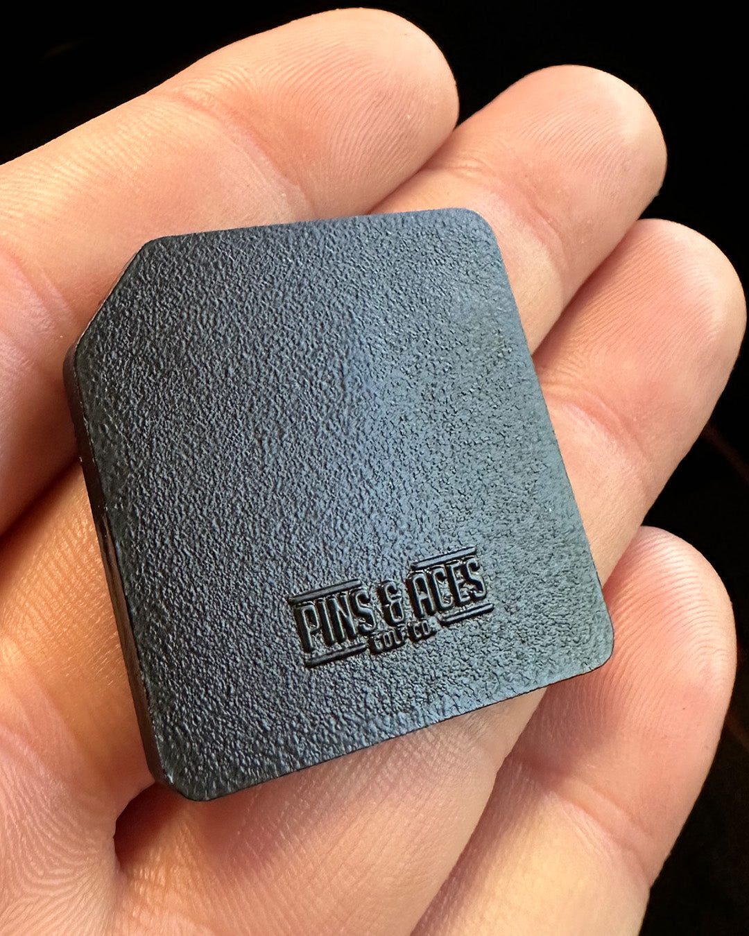 Floppy disc ball marker in a hand, featuring a textured black surface and the Pins and Aces logo. Perfect for golfers.