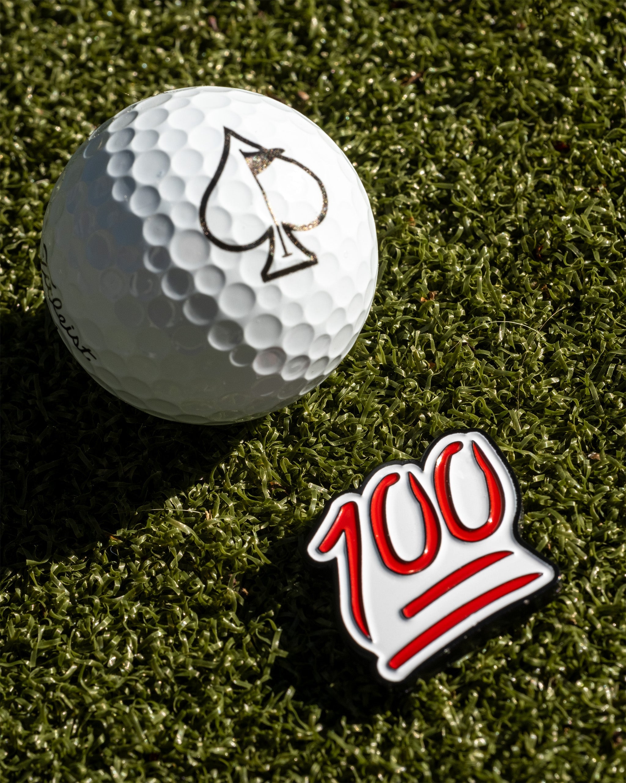 Bold golf ball marker featuring a vibrant "100" design next to a golf ball with a spade logo, set on green turf.