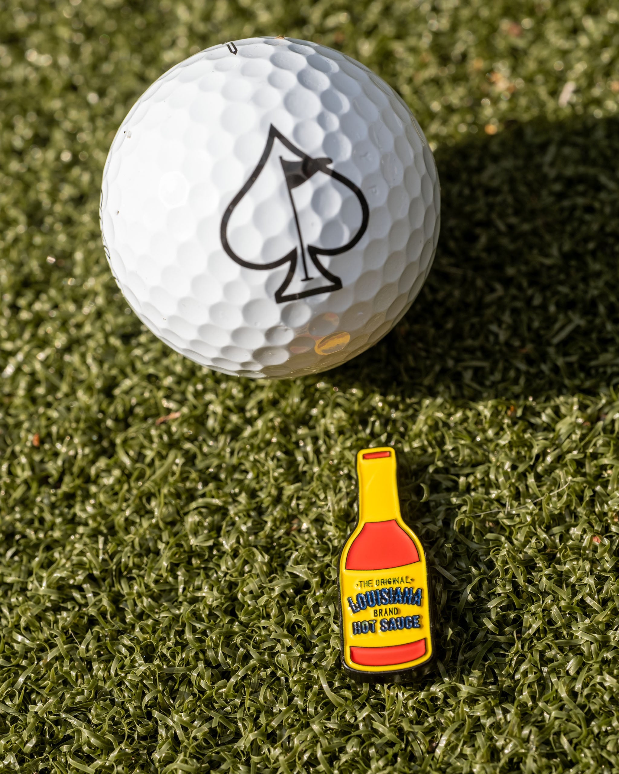 Louisiana Hot Sauce ball marker beside a golf ball with an ace of spades logo on a green turf background.