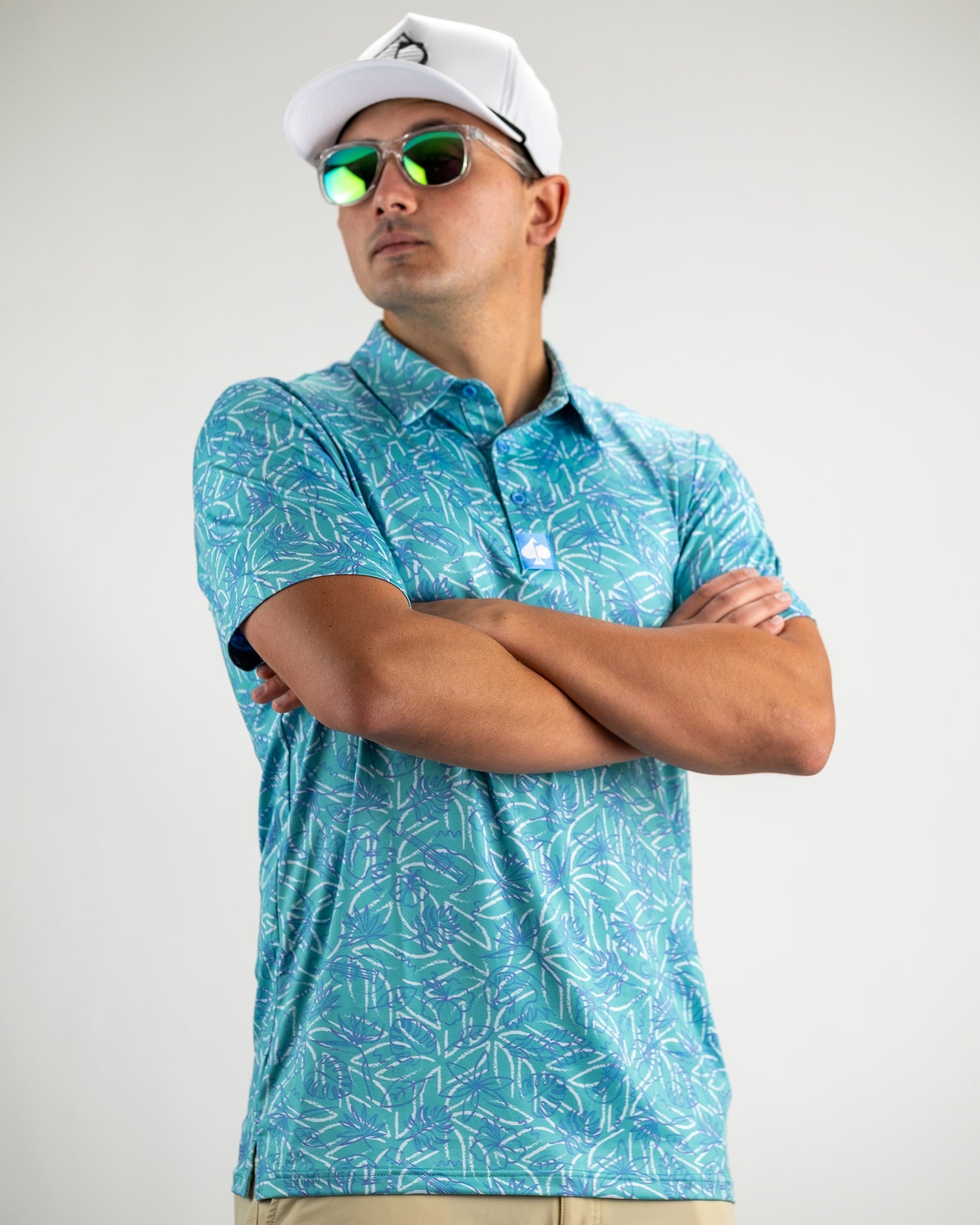 Model wearing a Margaritaville 5 O'Clock Ocean polo shirt with a tropical leaf pattern, sunglasses, and a white cap.