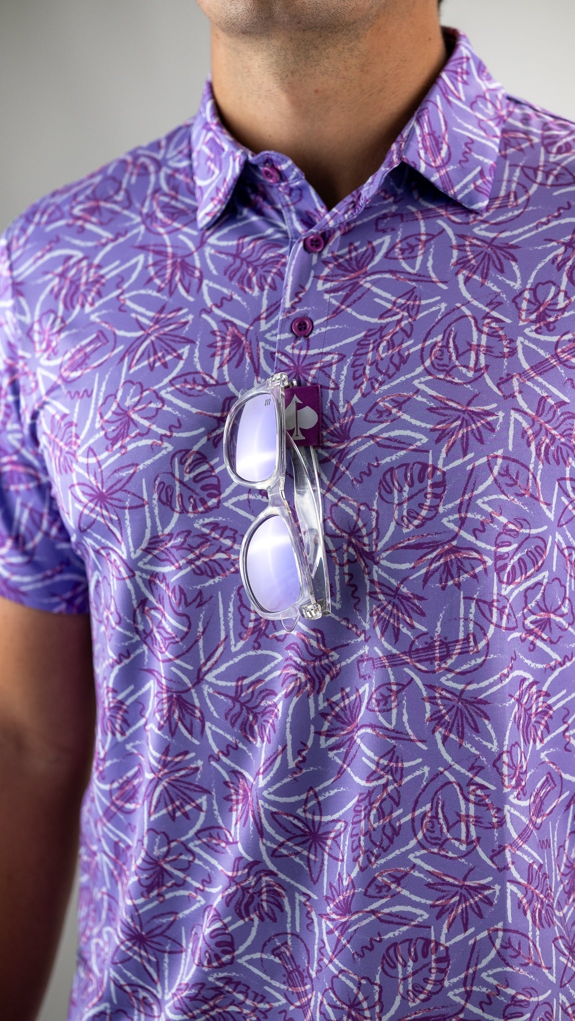 Men's Margaritaville 5 O'Clock Sunset polo shirt in purple with tropical print, featuring sunglasses draped over the collar.