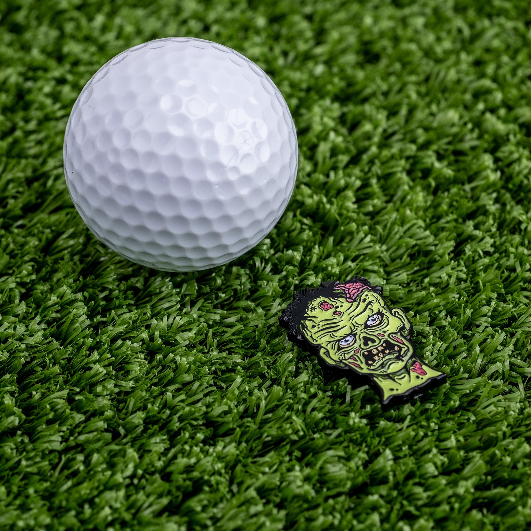 Bold Mutant Zombie ball marker next to a white golf ball on green turf, perfect for golfers wanting a unique accessory.