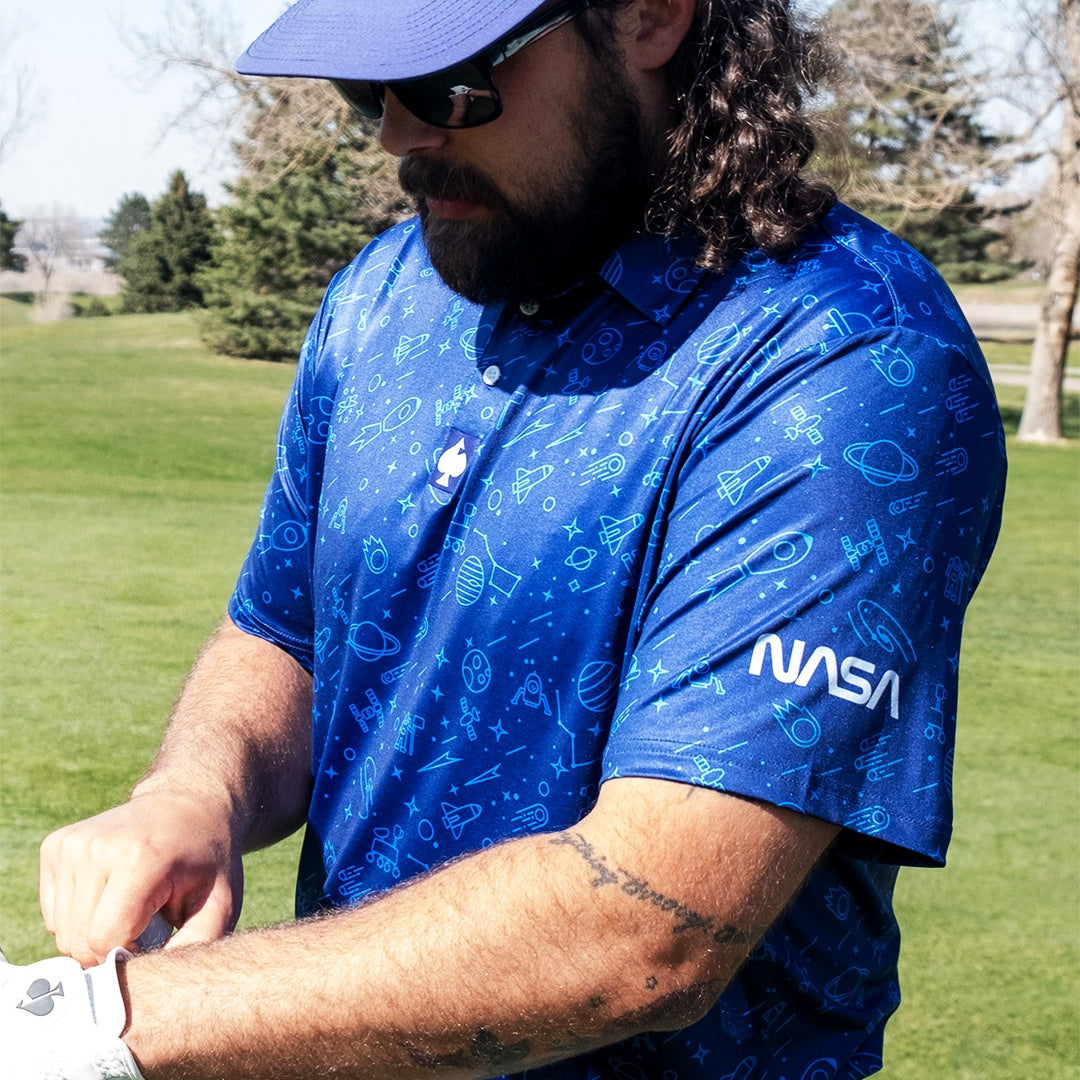 A golfer adjusts his glove while wearing a blue NASA polo featuring space-themed graphics, standing on a green golf course.