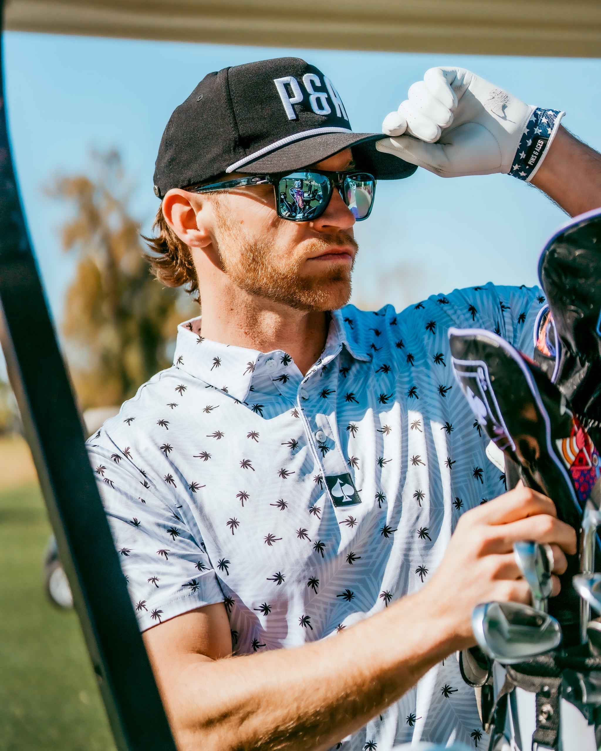 Stylish golfer adjusting his cap while wearing a palm-patterned polo shirt, showcasing bold golf fashion on the course.