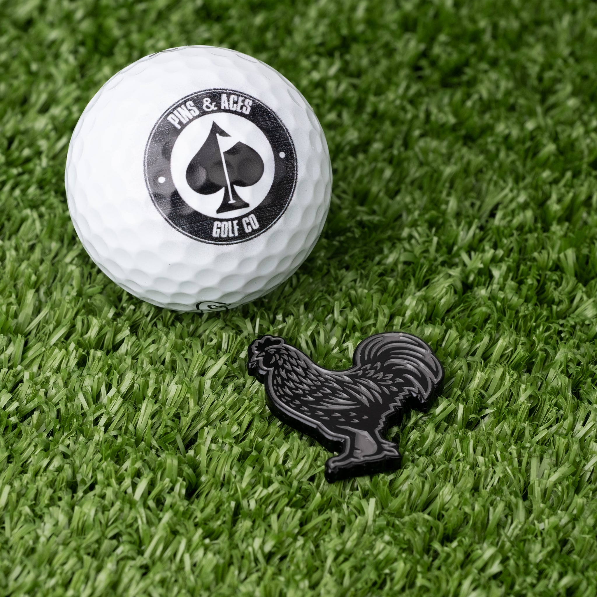 Black cock ball marker next to a golf ball on green turf, showcasing a unique design for bold golfers.