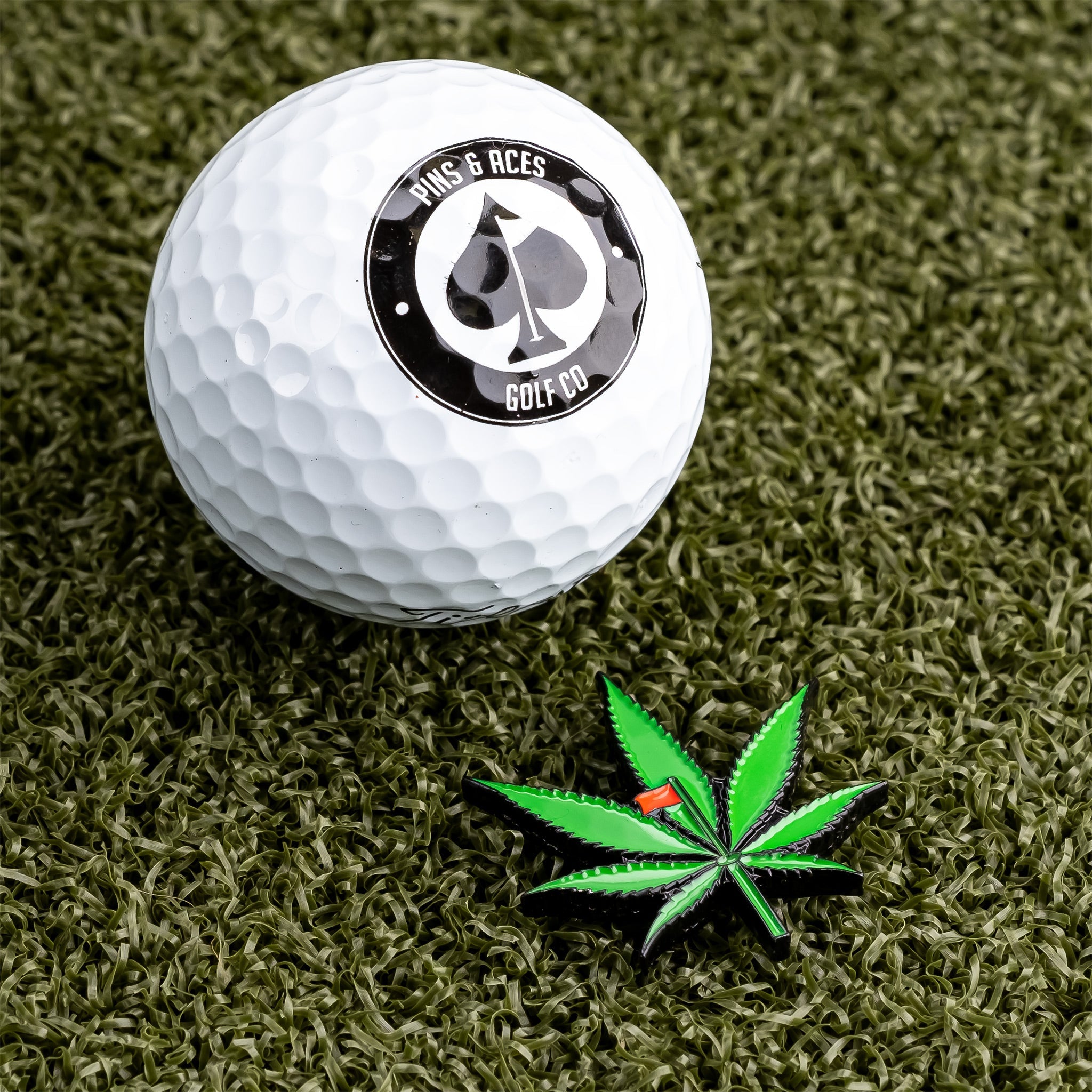 Pot leaf ball marker beside a golf ball featuring the Pins and Aces logo on a green turf background.
