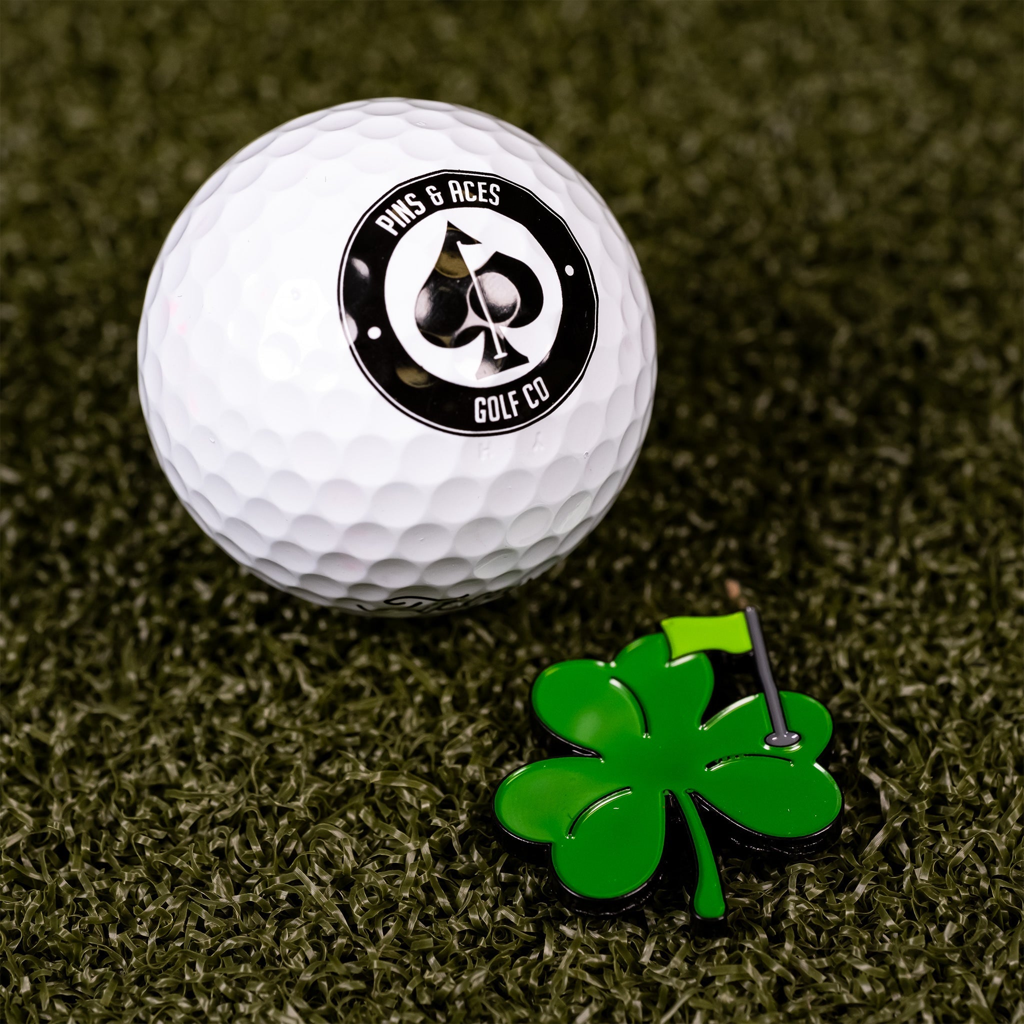 Shamrock ball marker beside a golf ball featuring the Pins and Aces logo, set on a green turf background.