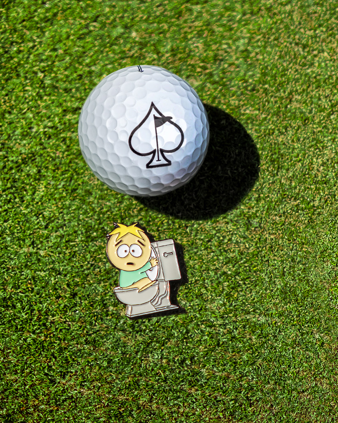South Park Butters ball marker next to a golf ball with an ace of spades logo on a green golf course.