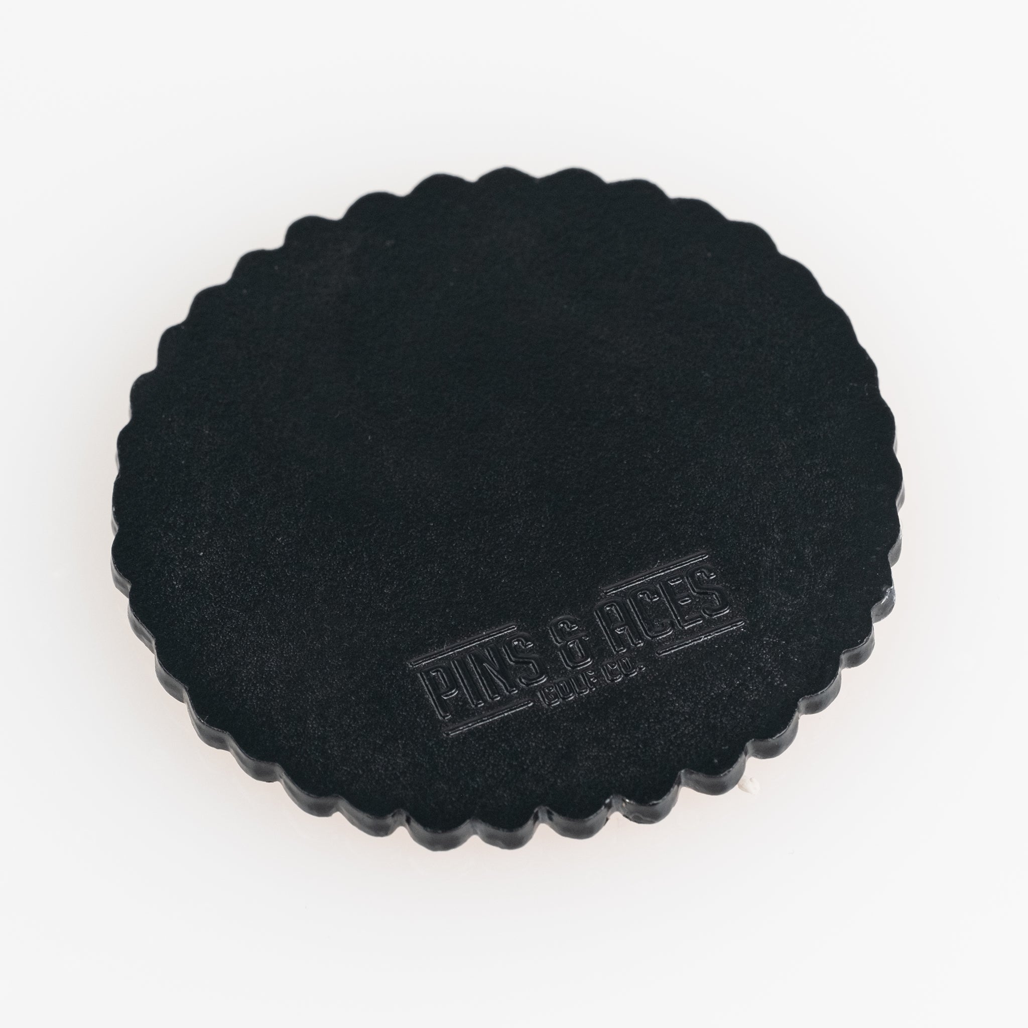 Black circular ball marker with a scalloped edge, featuring the embossed logo of Pins and Aces. Ideal for golfers.
