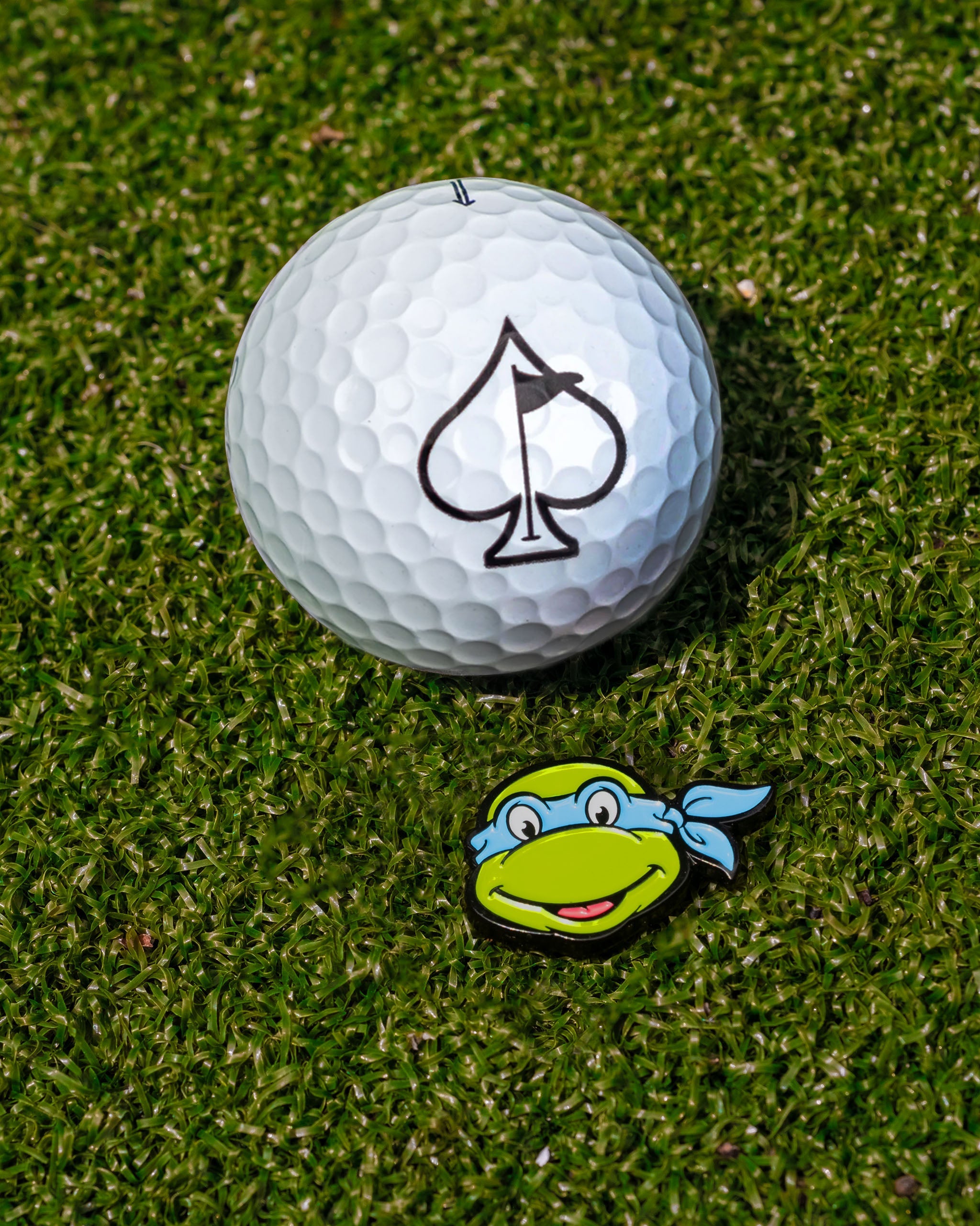 Leonardo ball marker featuring a green turtle head with a blue bandana next to a golf ball on grass.