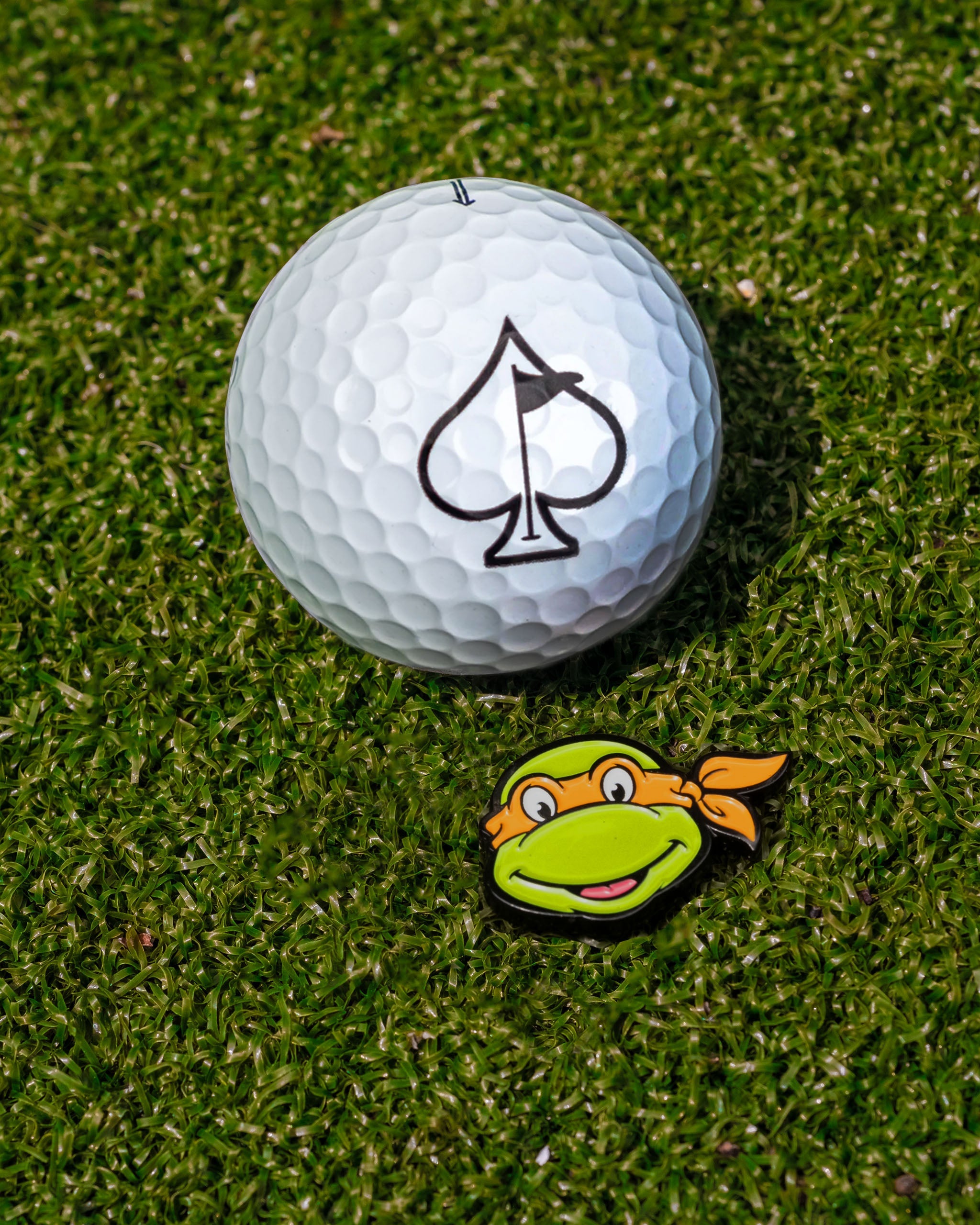 TMNT Michelangelo ball marker beside a golf ball with an ace of spades logo, set on green grass.