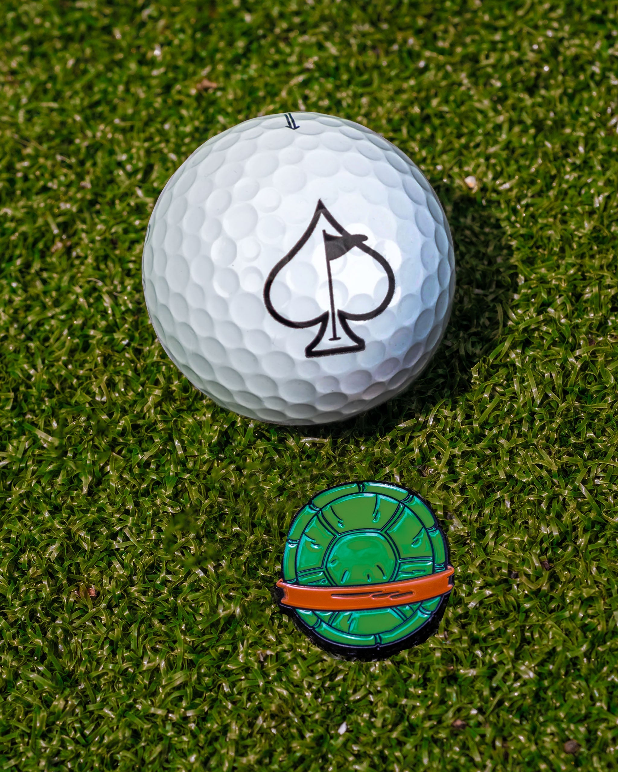 TMNT Shell Shock Ball Marker beside a golf ball with an ace of spades logo on a grassy surface.