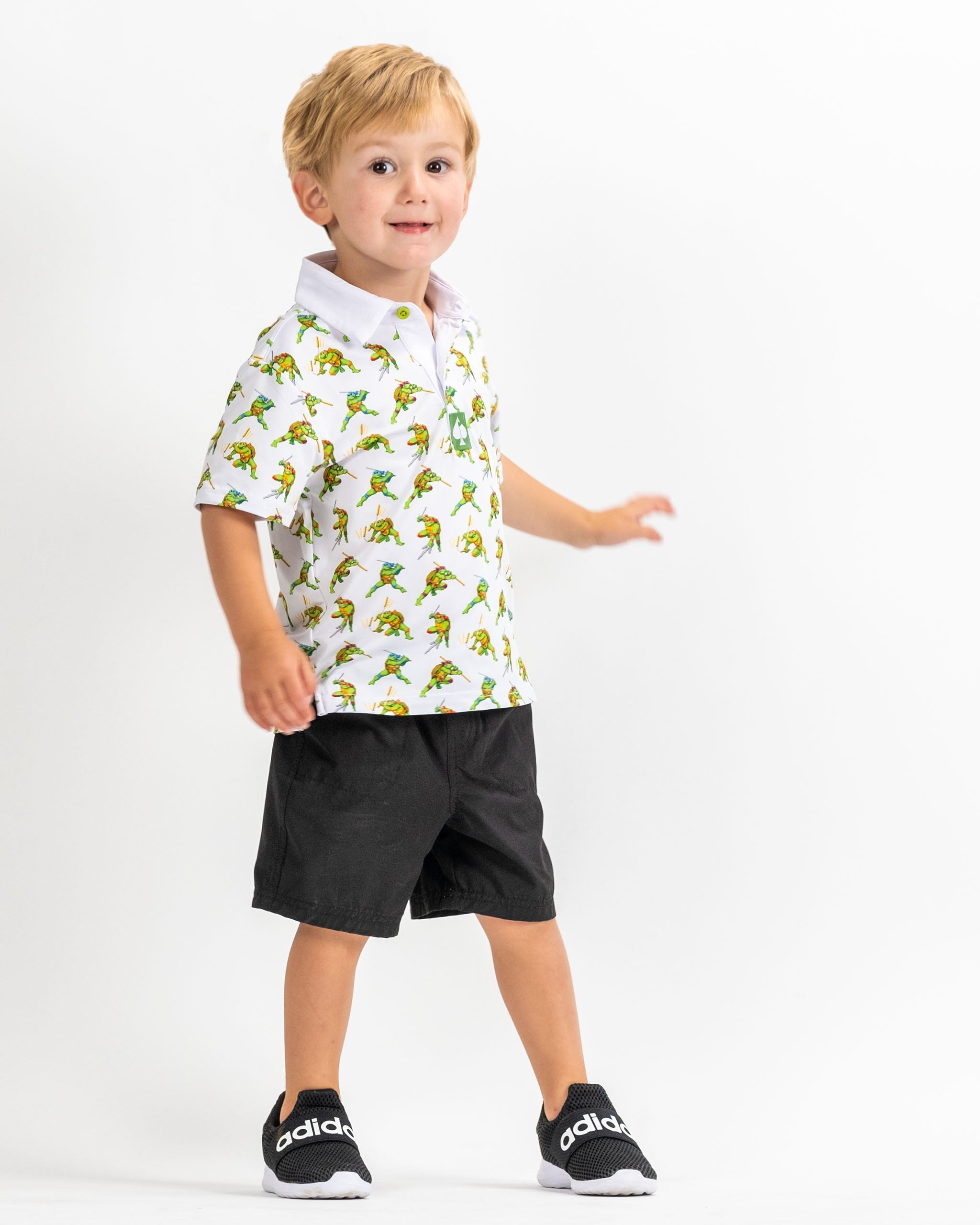 Youth in a TMNT-themed polo shirt featuring colorful 8-bit turtle graphics, paired with black shorts and sneakers.