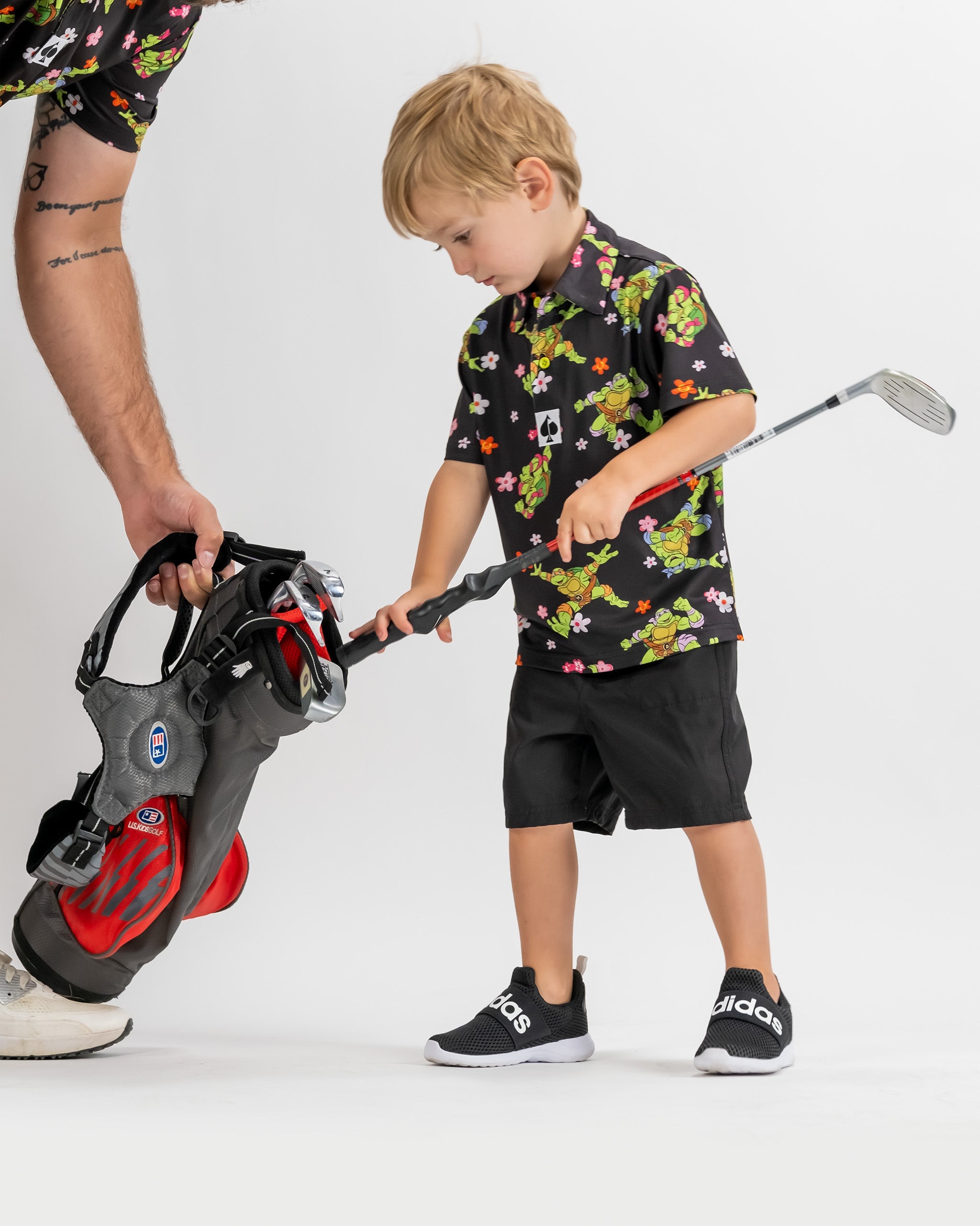 Youth in a vibrant TMNT floral golf shirt and black shorts, preparing to play golf with a club and bag.