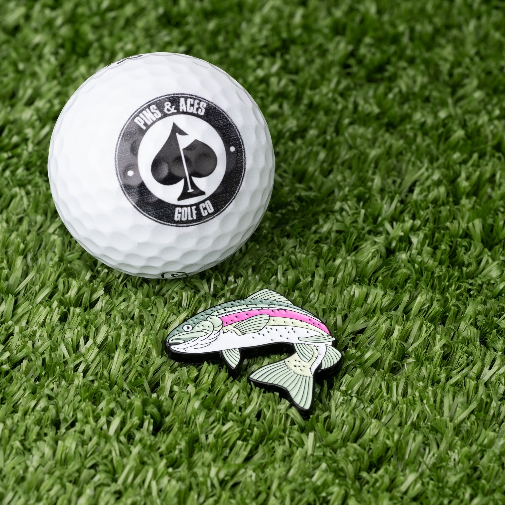 Trout ball marker beside a golf ball on green turf, featuring a colorful fish design for stylish golfing.