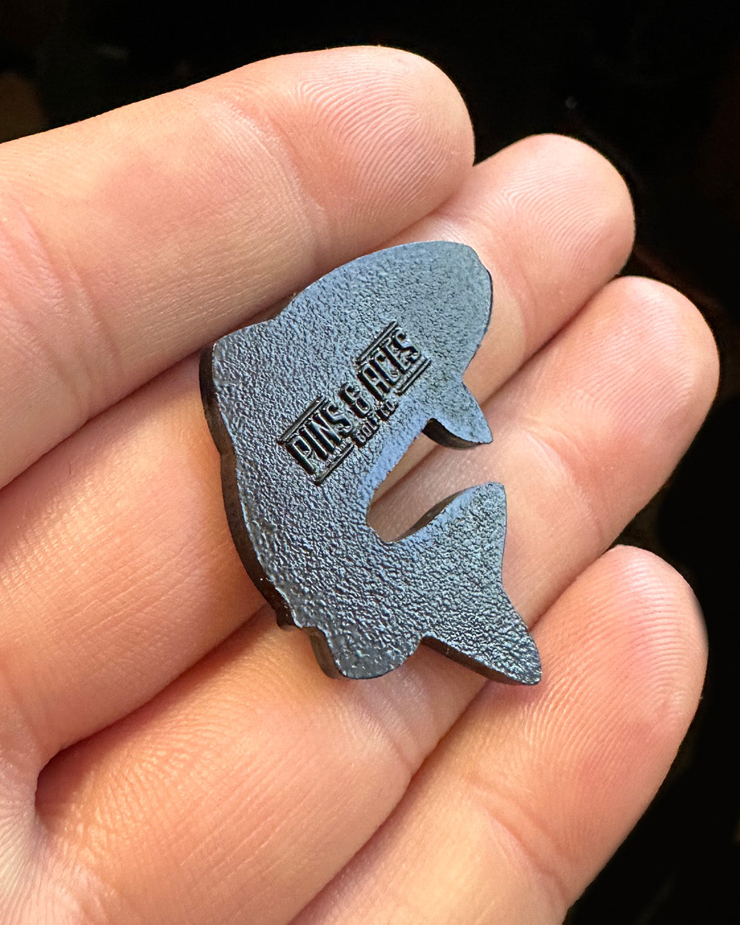 Trout-shaped ball marker held in a hand, featuring the Pins and Aces logo for stylish golfing.