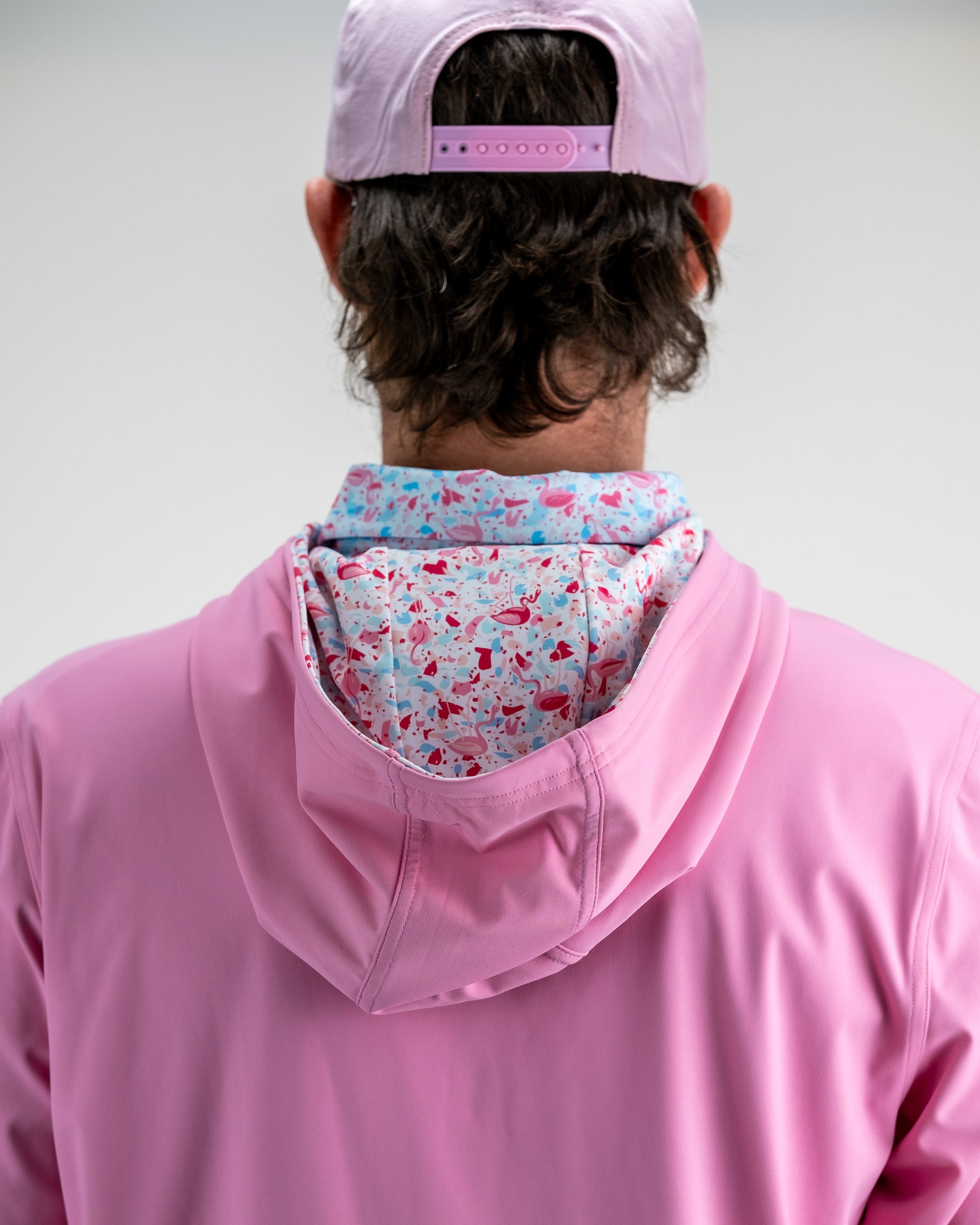 Back view of a golfer wearing a pink cap, pink jacket, and a colorful flamingo-patterned hoodie, perfect for bold golf style.