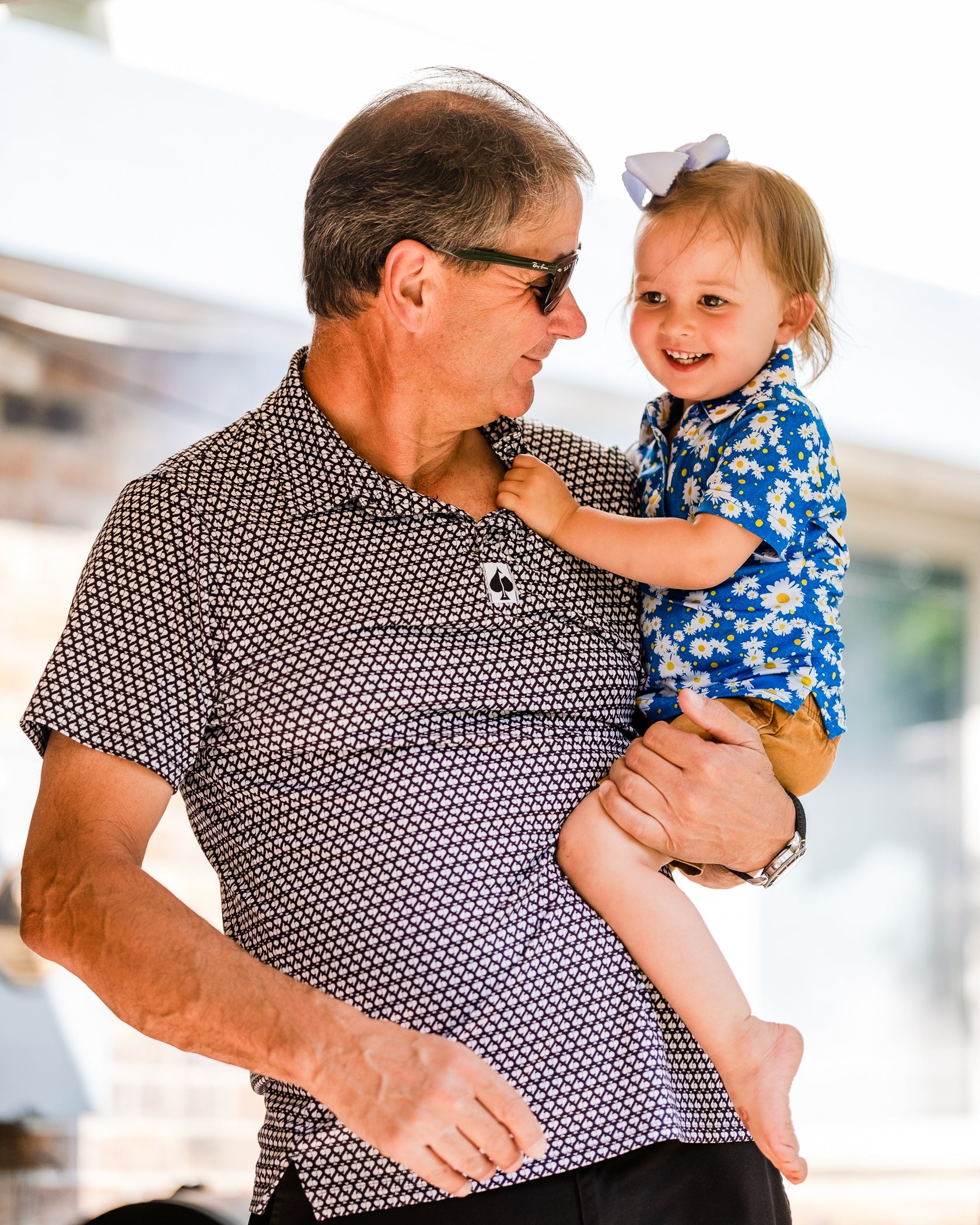 A man holds a smiling young girl wearing a blue daisies polo shirt, enjoying a sunny day outdoors.