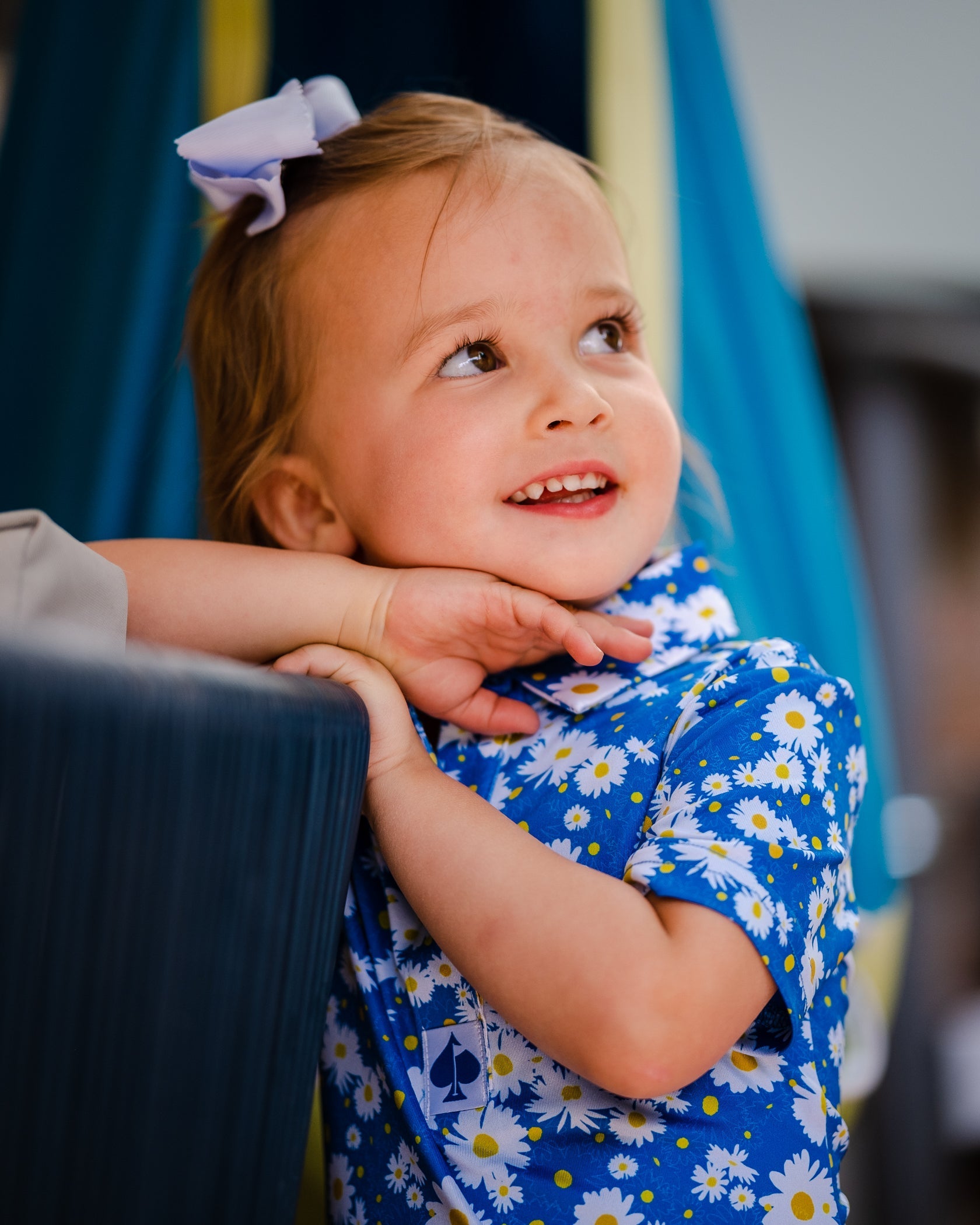 Youth girls' blue polo shirt with a daisy pattern, worn by a smiling girl with a bow in her hair, posing playfully.