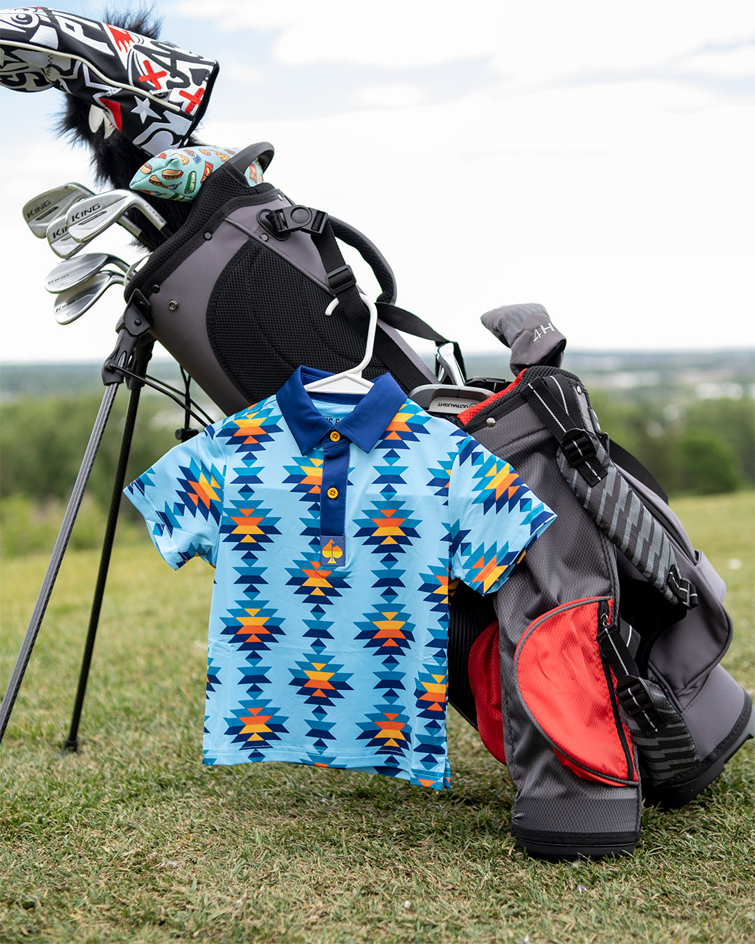 Youth Southwest Sunset Polo hanging on a golf bag, featuring a vibrant blue and orange geometric pattern, perfect for young golfers.