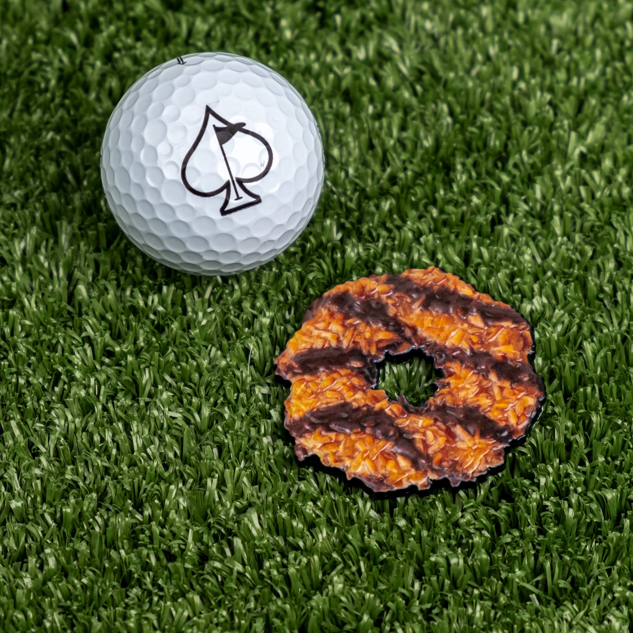 Coconut fudge cookie ball marker next to a golf ball on green turf, showcasing a fun and unique golf accessory.