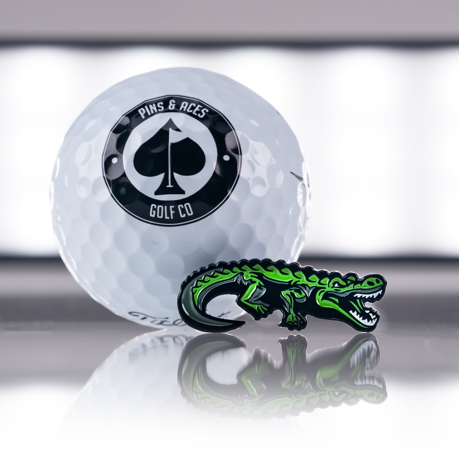Crocodile ball marker in vibrant green next to a white golf ball featuring the Pins and Aces logo.