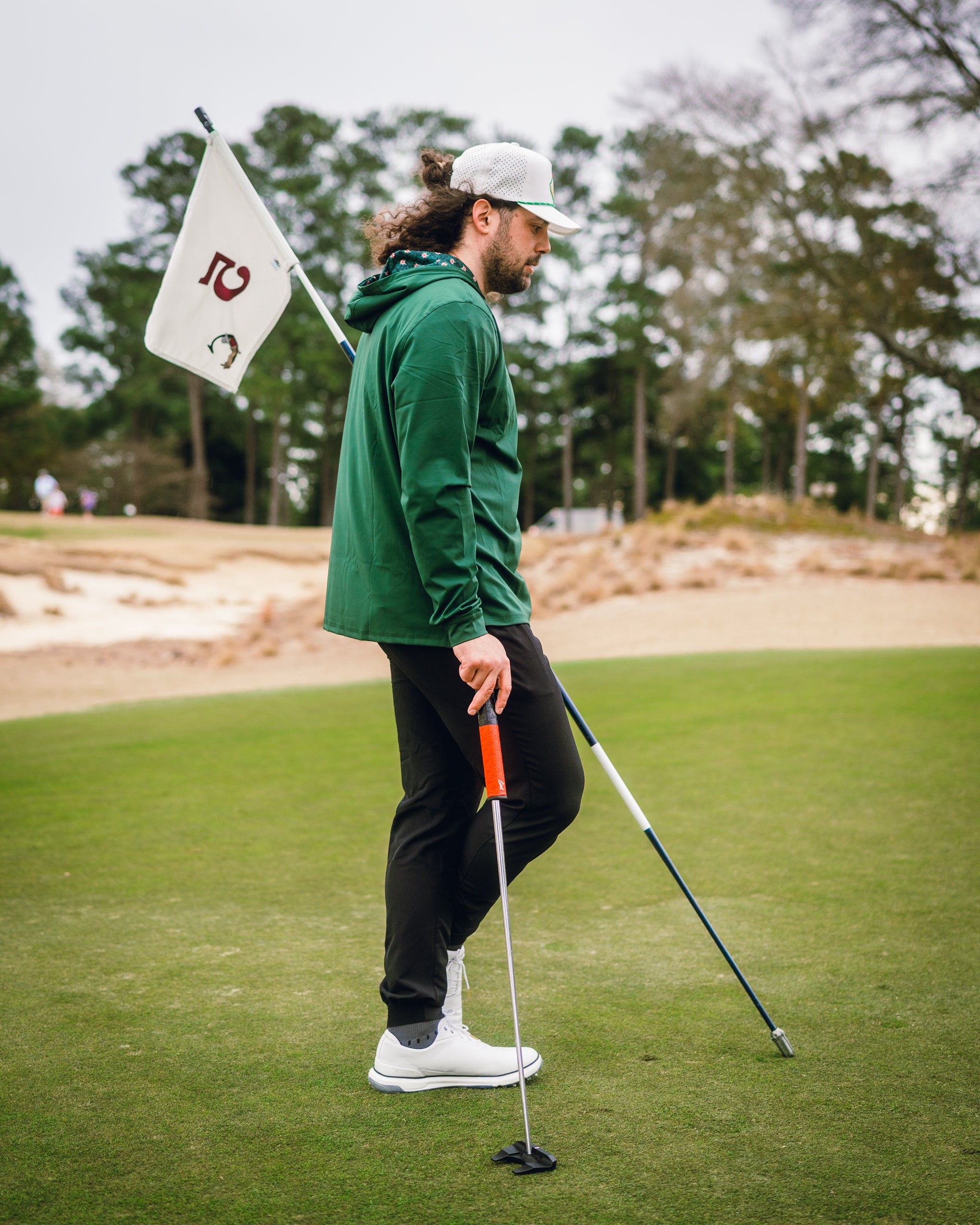 A golfer wearing a dark azalea performance hoodie walks on the course, holding a flag and a putter, showcasing stylish golf apparel.