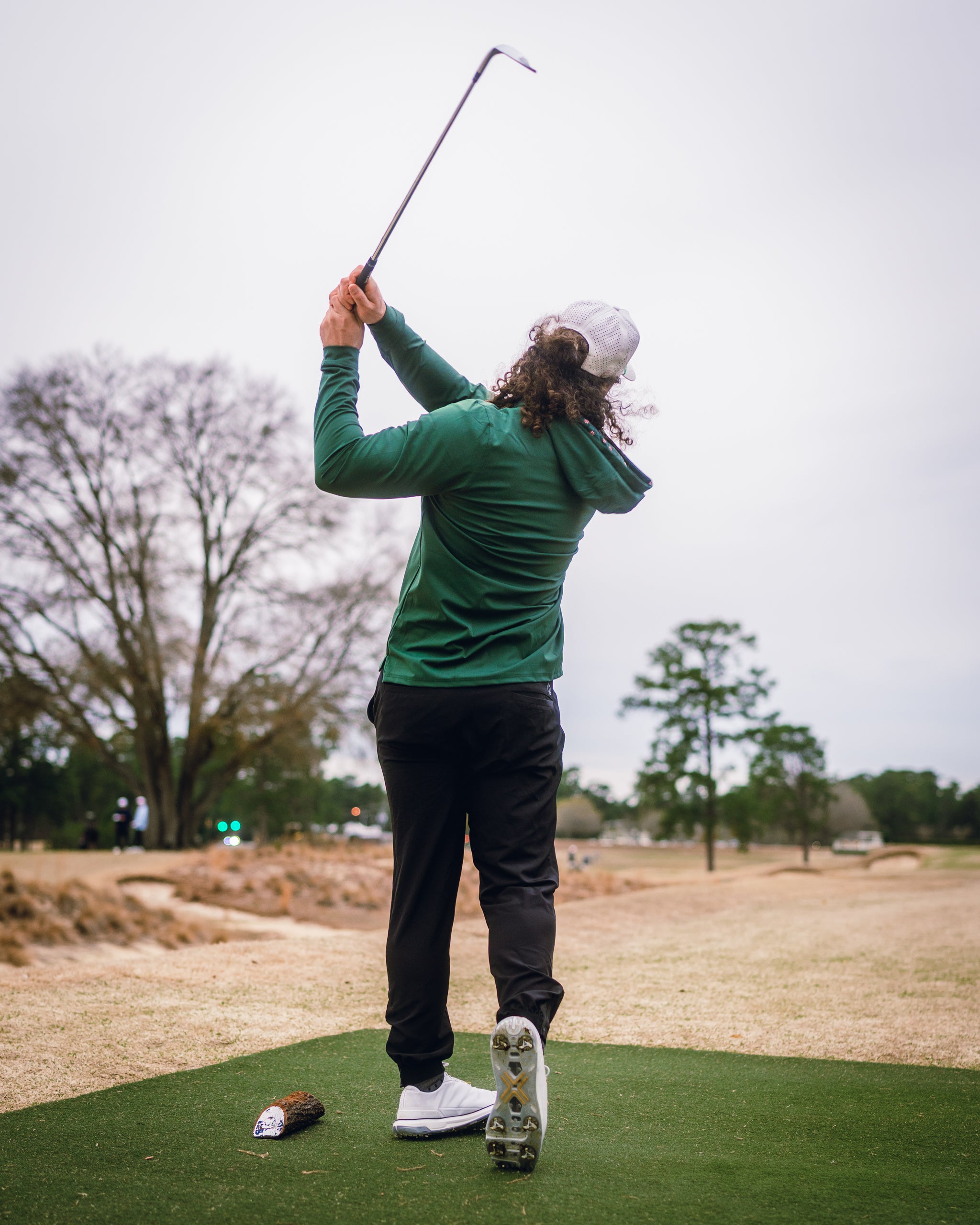 A golfer swings a club on the course, wearing a dark azalea performance hoodie and black pants, showcasing stylish golf apparel.