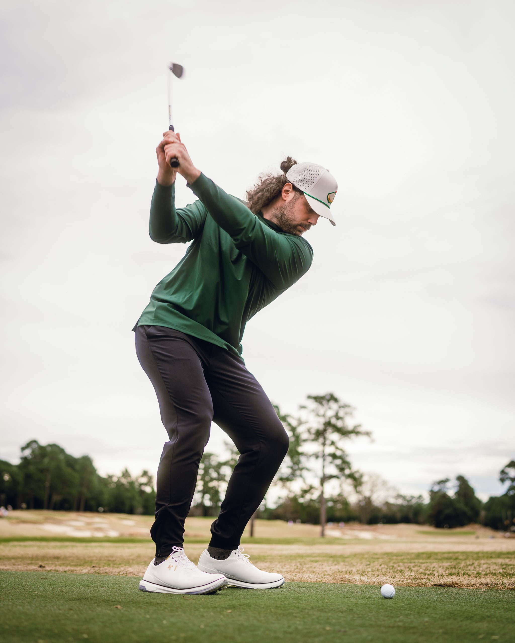 A golfer swings a club while wearing a dark azalea performance hoodie, showcasing a stylish and functional golf outfit.