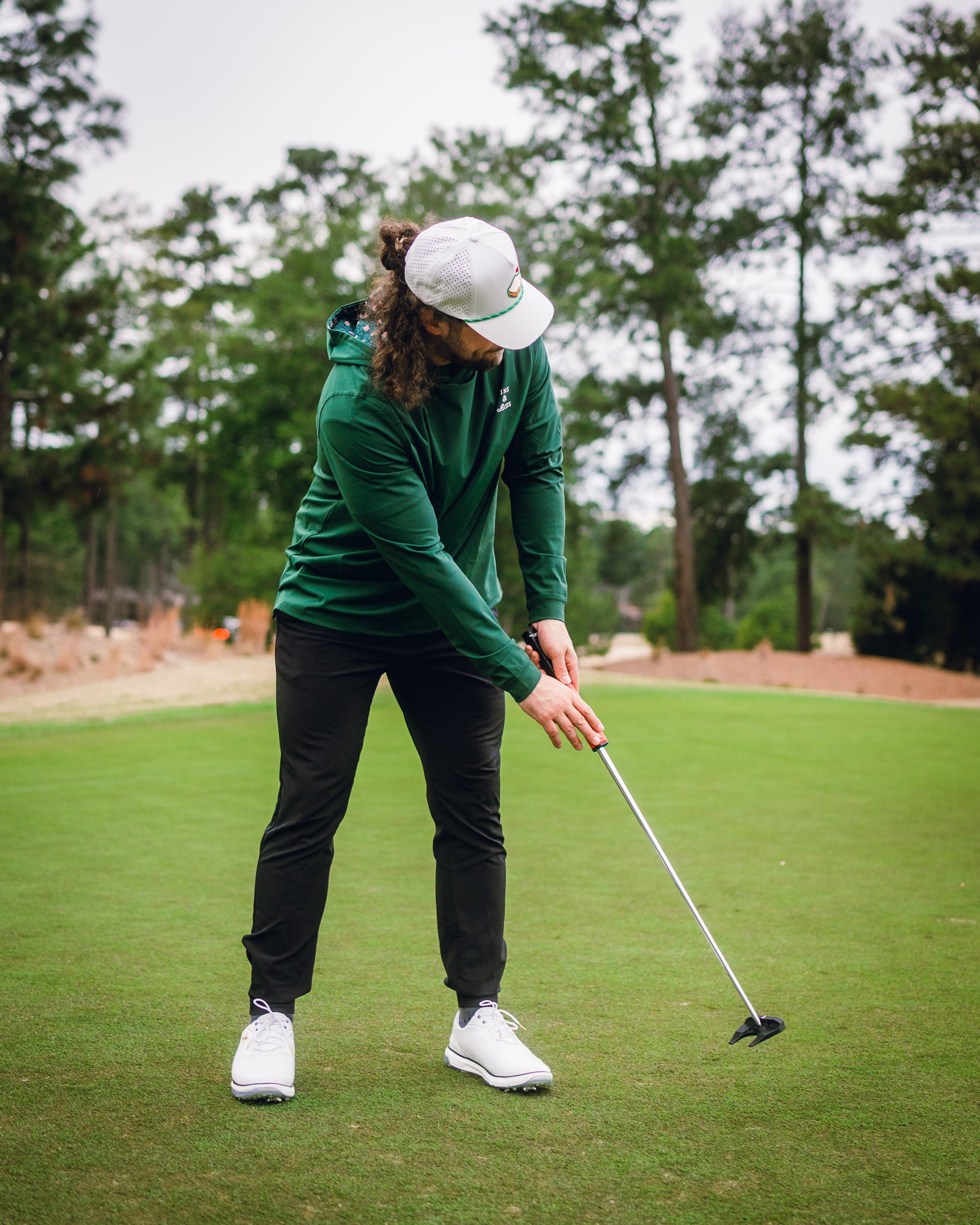 A golfer wearing a dark azalea performance hoodie and black pants prepares to putt on a lush green course.