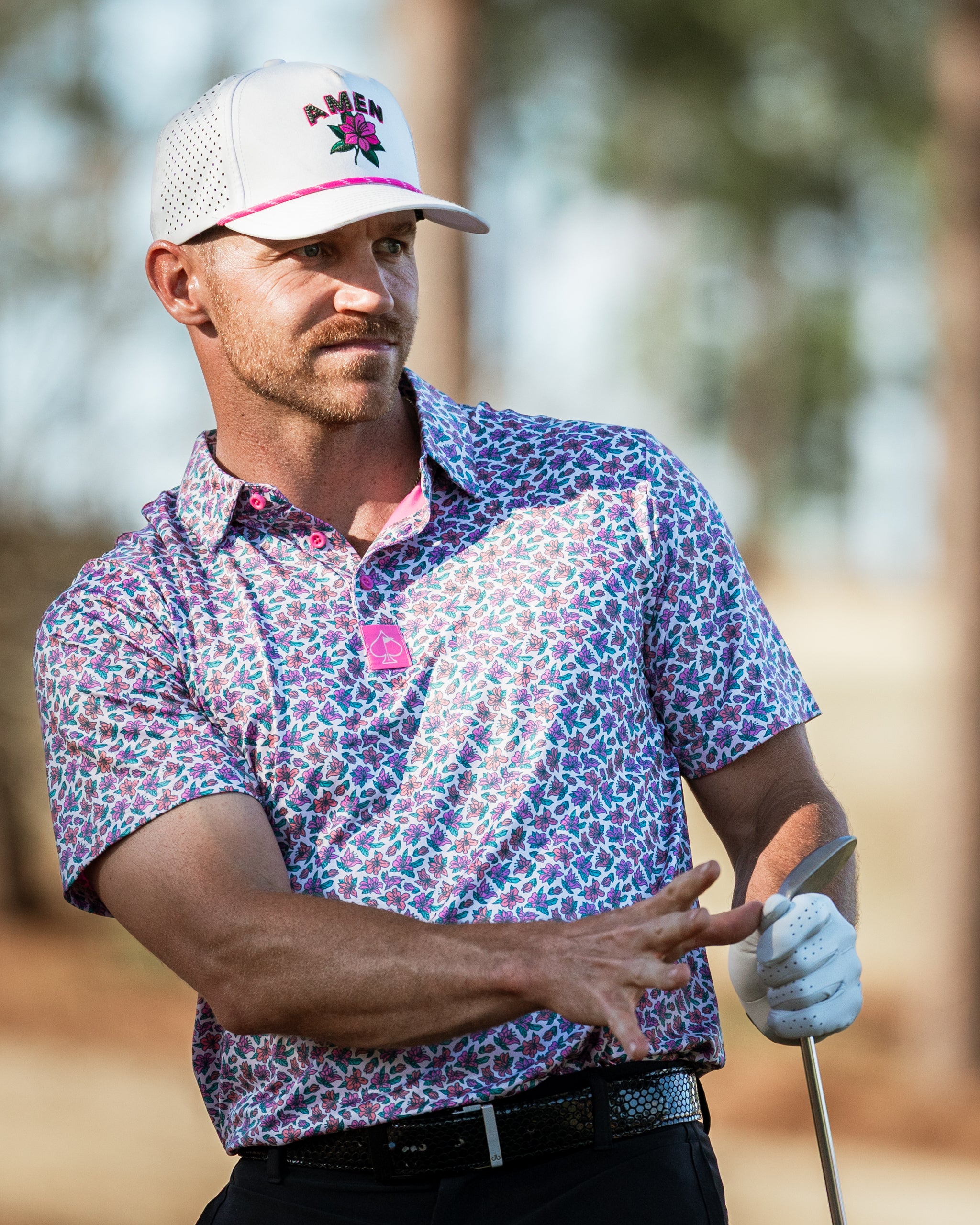 Man wearing a floral-patterned golf shirt and a white cap, preparing to swing a golf club on the course.