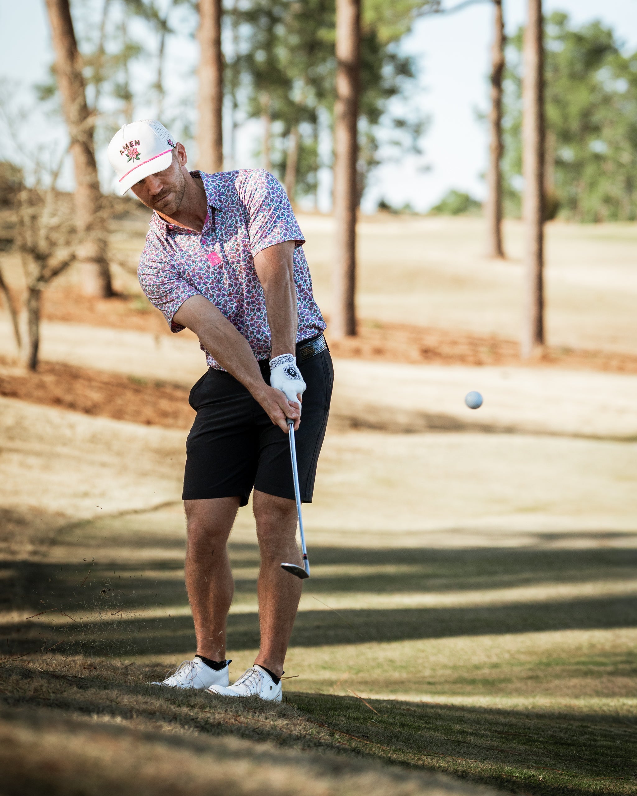 A golfer in a light azalea-patterned shirt and black shorts swings a club on a sunny course, with trees in the background.