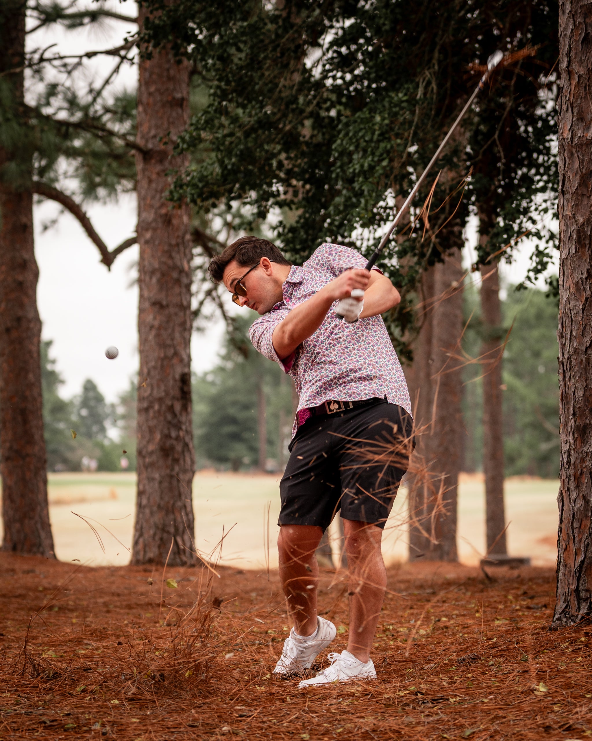 A golfer in a light azalea patterned shirt swings a club in a wooded area, with pine needles flying around him.