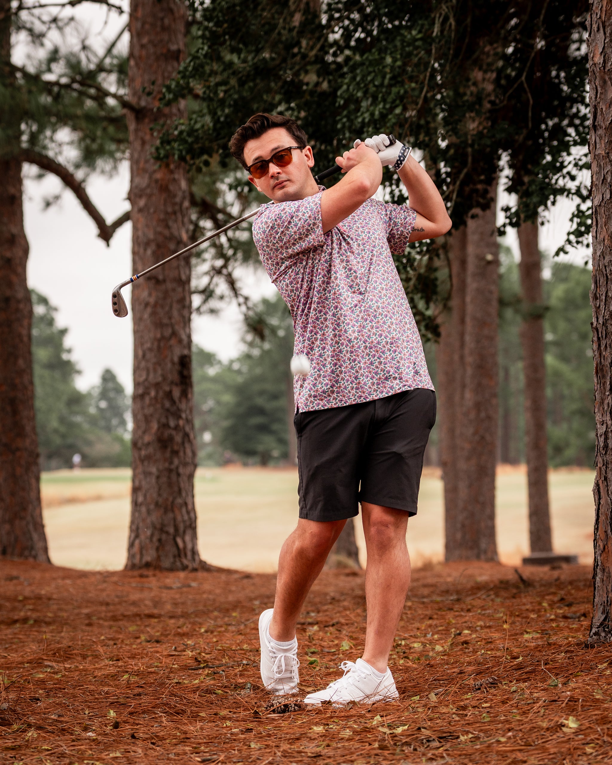 A golfer swings a club in a floral light azalea shirt and shorts, surrounded by tall trees on a golf course.