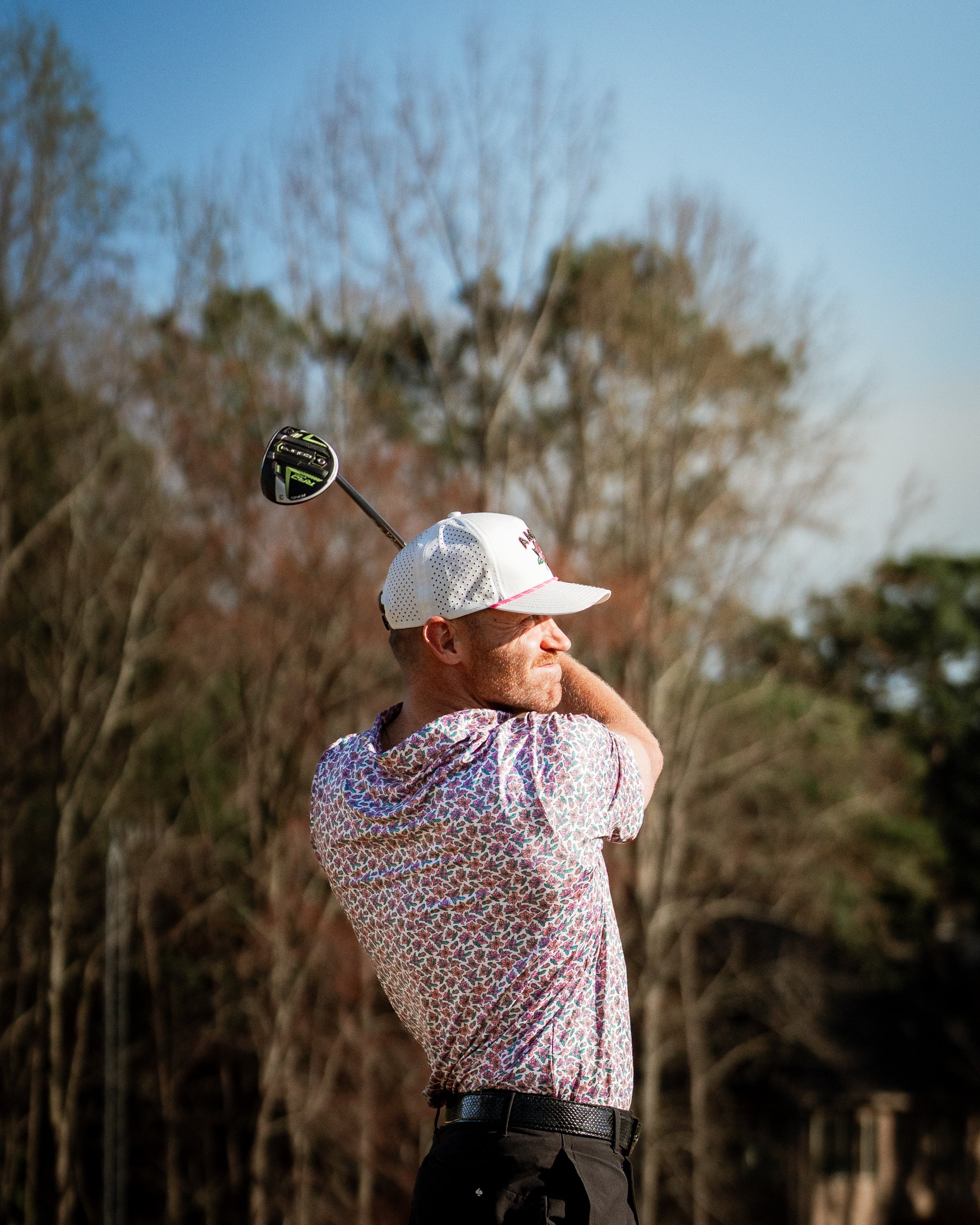 A golfer swings a driver while wearing a light azalea-patterned shirt, showcasing bold golf fashion on the course.