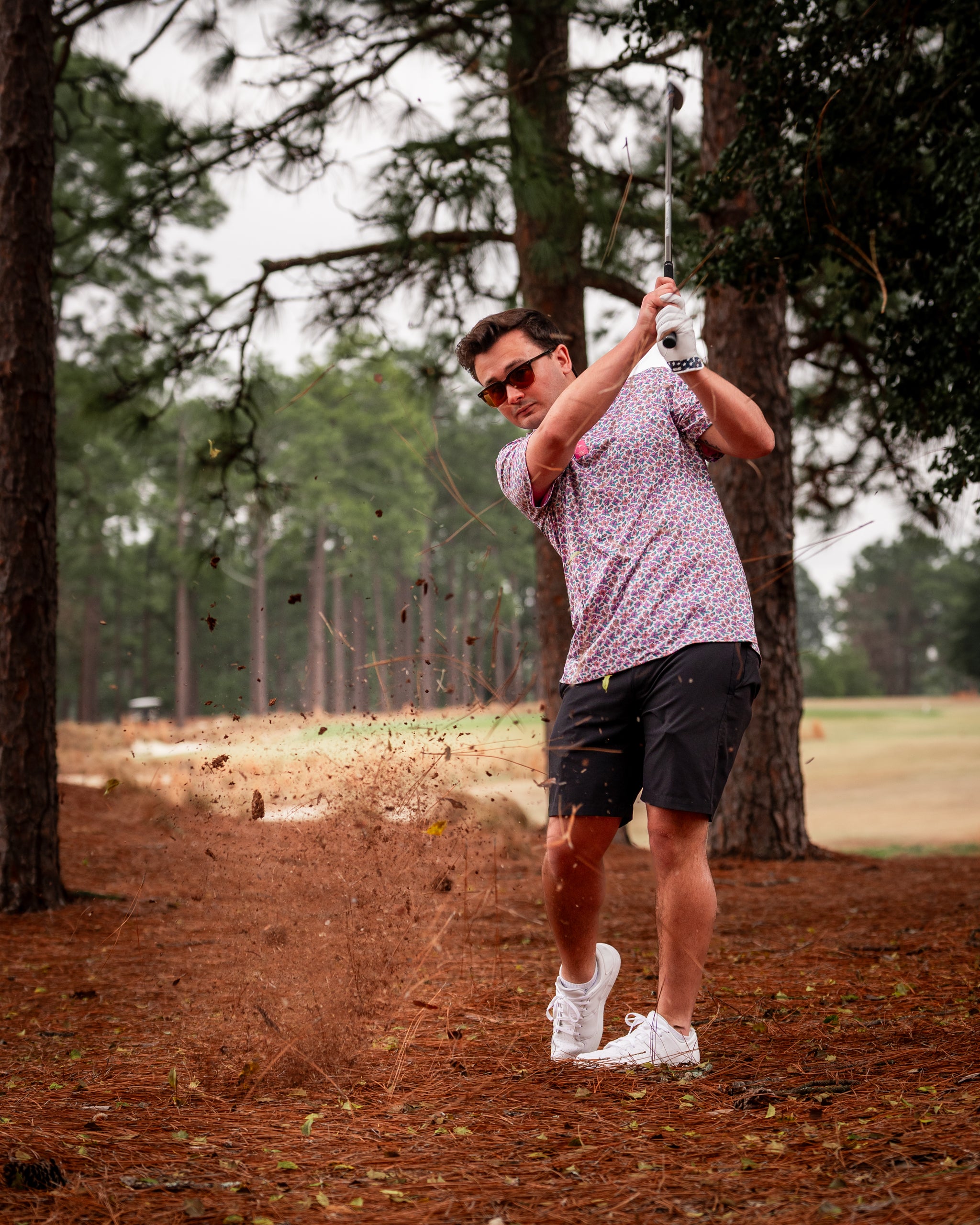 A golfer in a light azalea patterned shirt swings a club, sending dirt flying in a wooded area on the course.