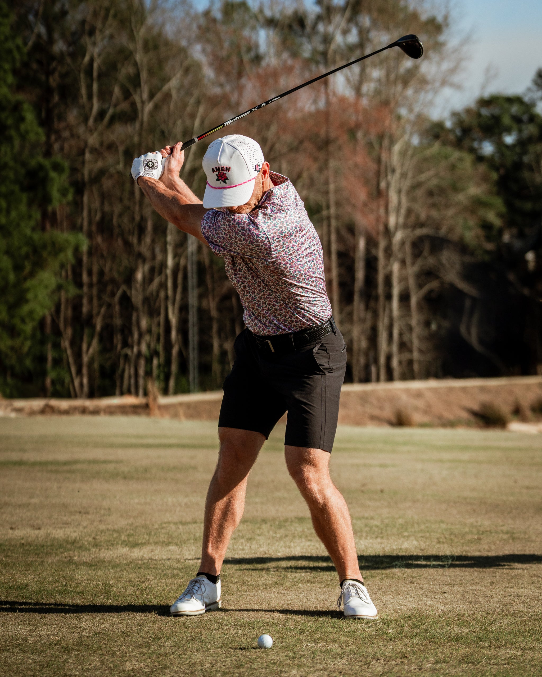 A golfer in a light azalea patterned shirt and cap swings a driver on a sunny course, showcasing bold golf style.