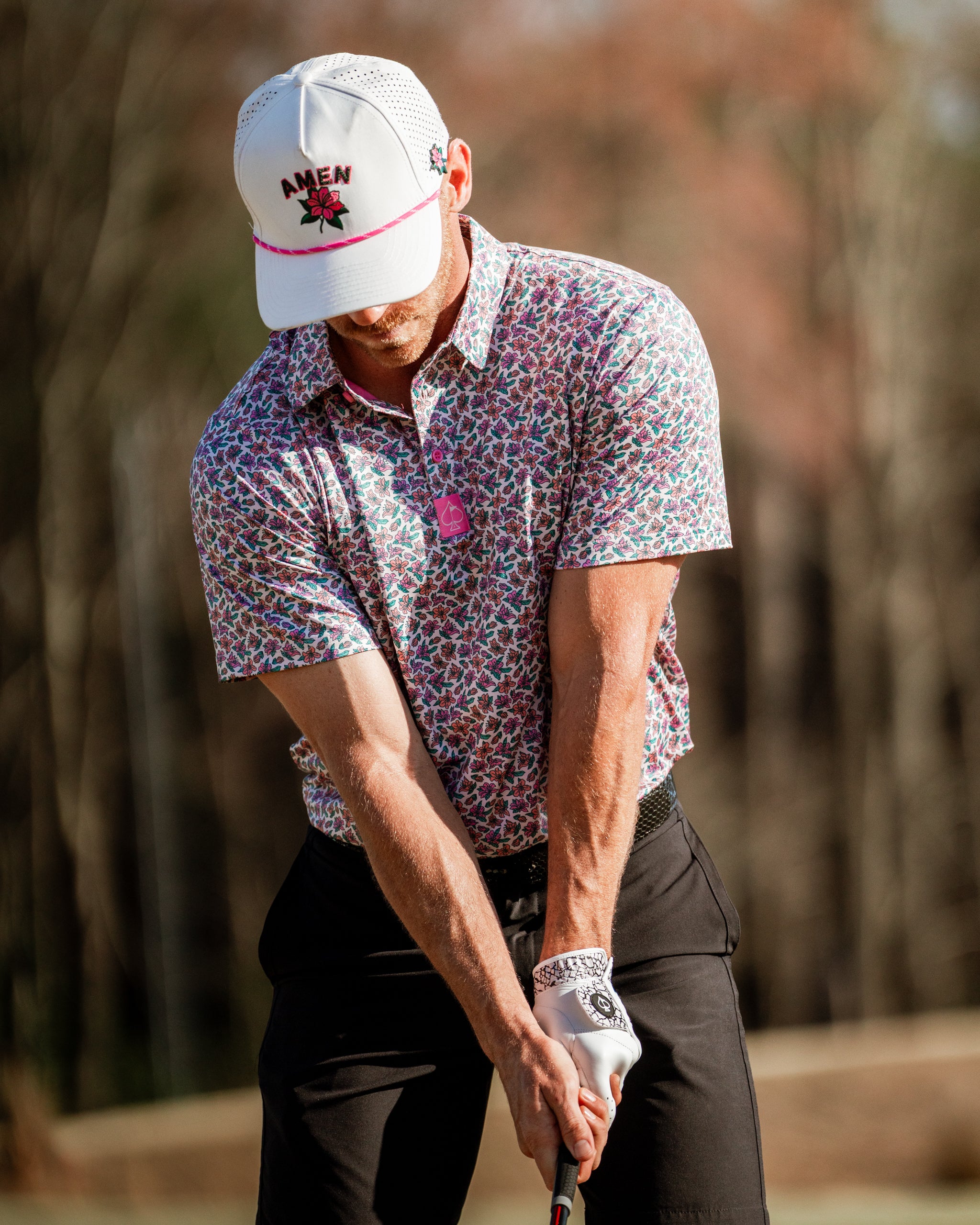 A golfer in a light azalea-patterned shirt and a white cap prepares to swing on the course, showcasing bold golf style.