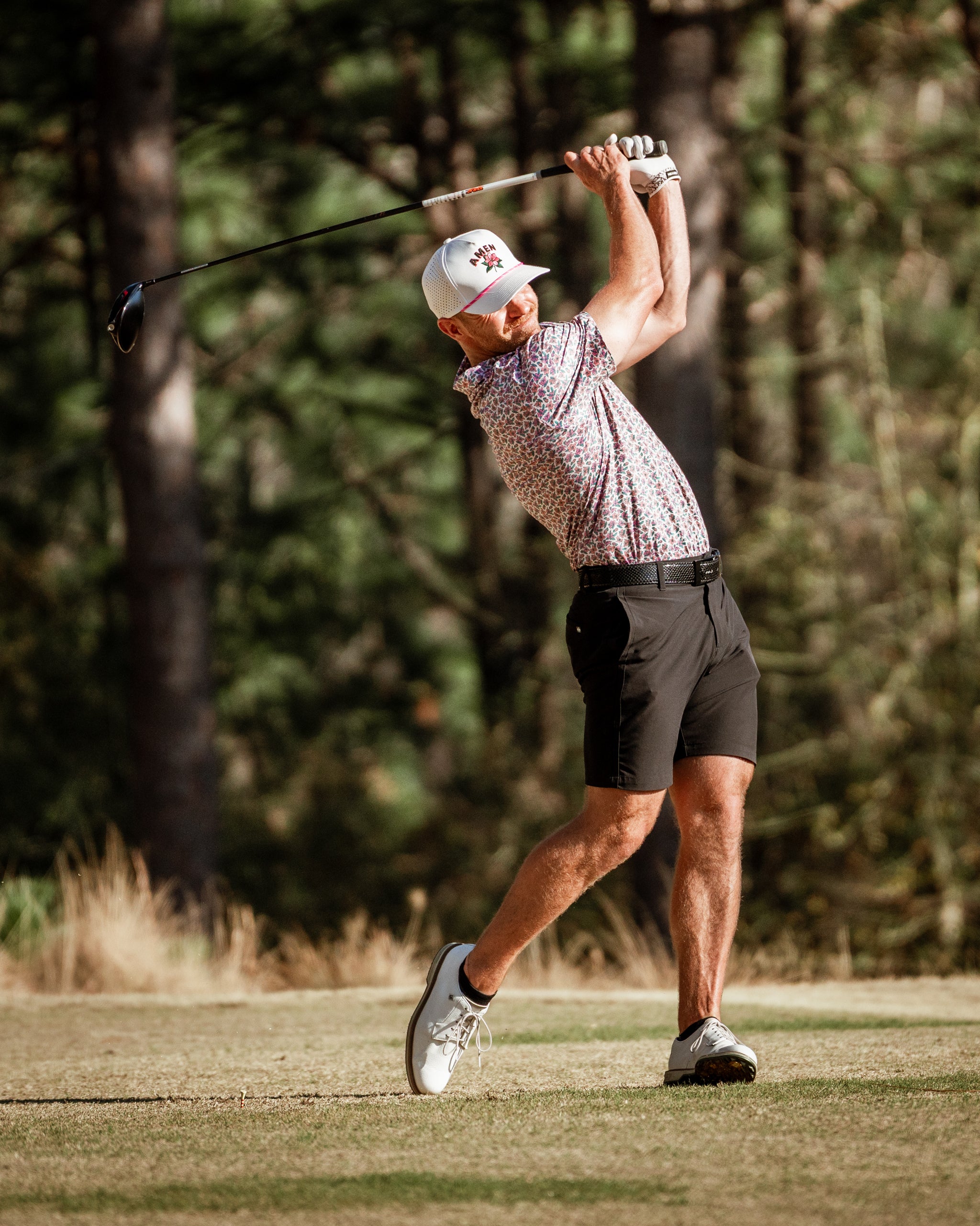 A golfer swings a club on the course, wearing a light azalea-patterned shirt and black shorts, showcasing bold golf style.