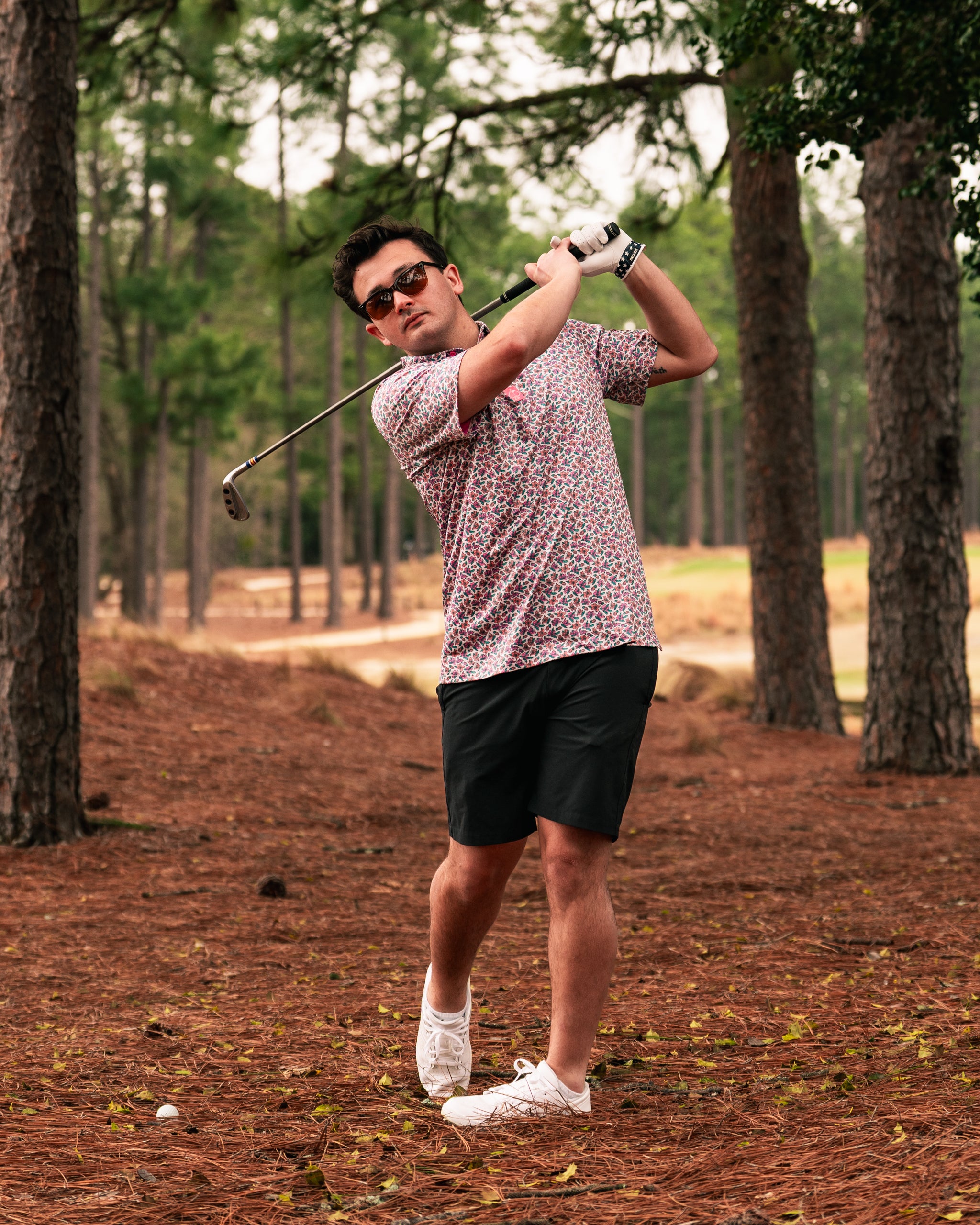 A golfer swings a club in a floral-patterned polo shirt and shorts, surrounded by tall trees on a golf course.