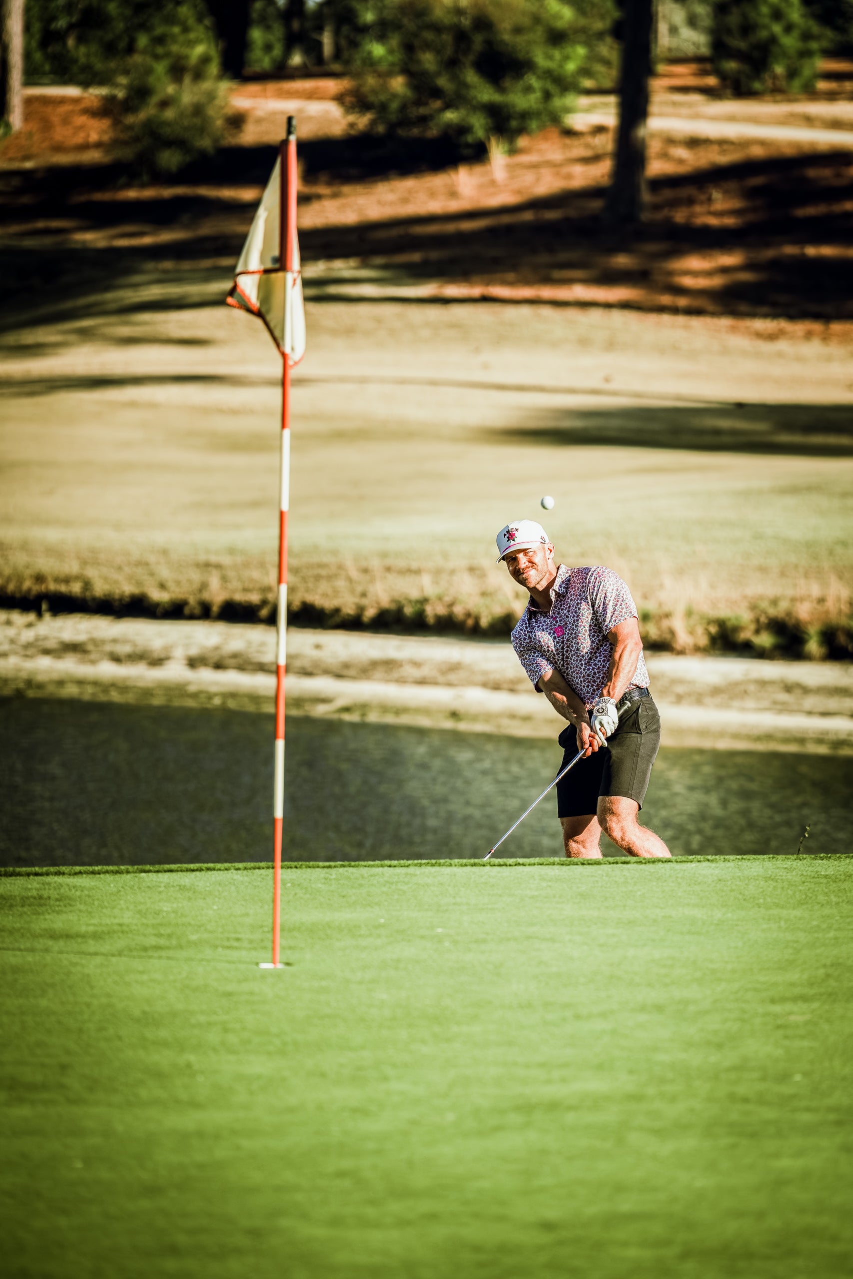 Golfer in a light azalea shirt chips onto the green near a flag, with a scenic water hazard in the background.