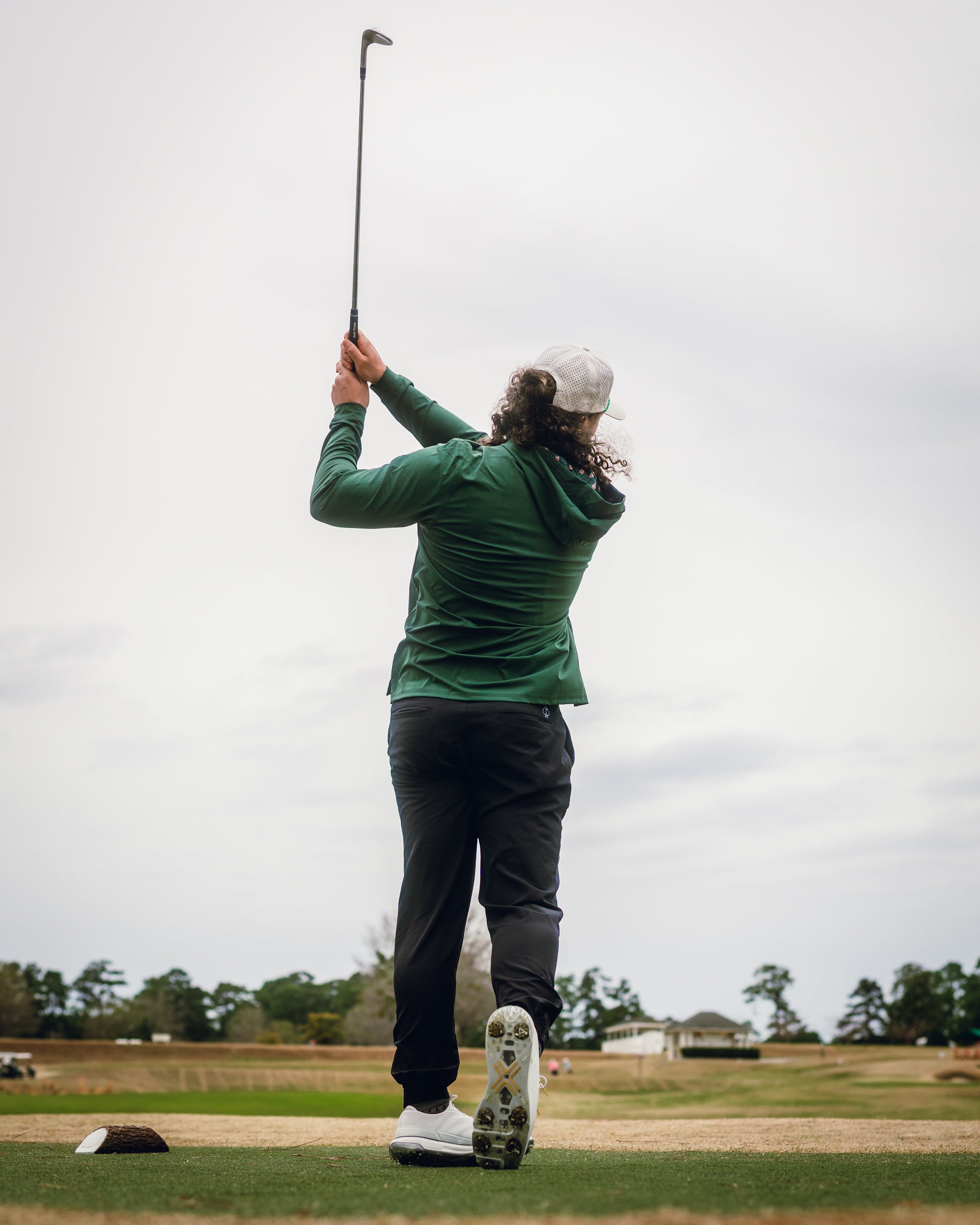 A golfer swings a club while wearing a dark azalea performance hoodie, showcasing stylish golf apparel on the course.