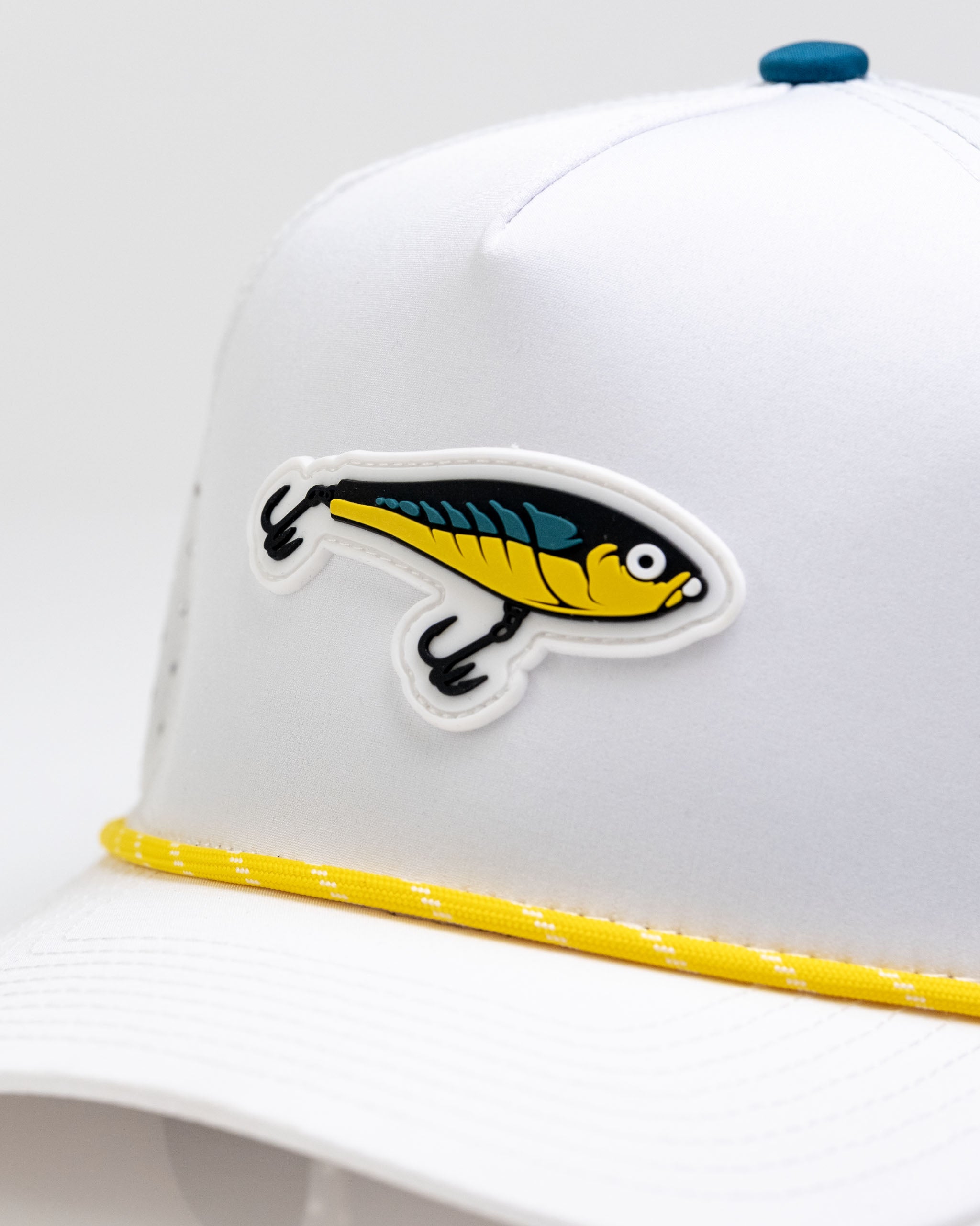 White perforated rope hat featuring a colorful fishing lure design and a yellow accent band. Perfect for fishing enthusiasts.
