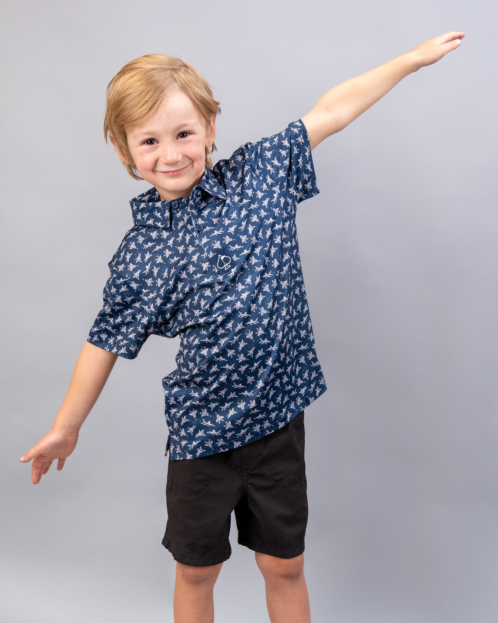 Young boy smiling and posing with arms outstretched, wearing a navy blue shirt with a bird pattern and black shorts.