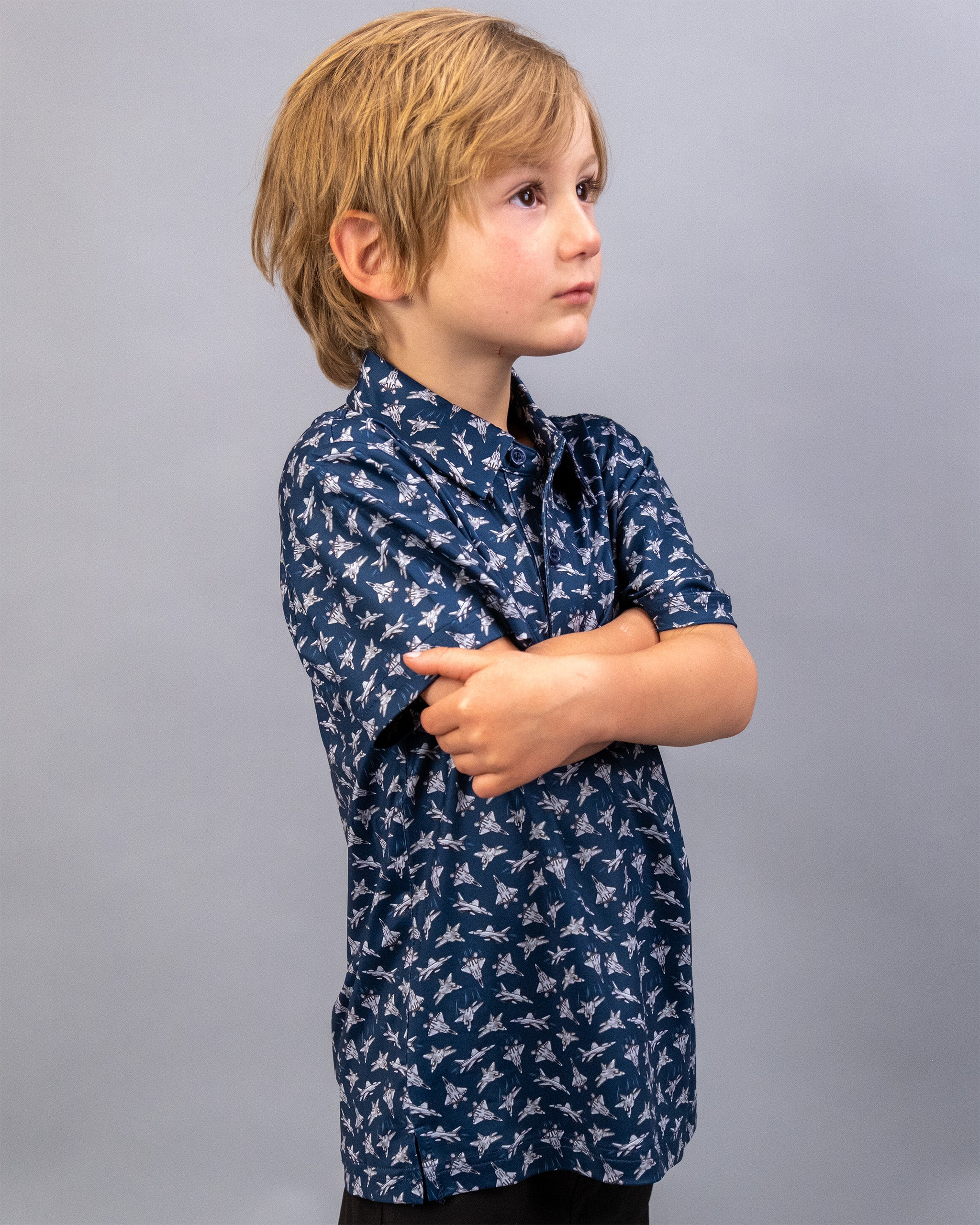 A young boy with light brown hair wearing a navy blue shirt with a white bird pattern, standing with arms crossed against a plain gray background.